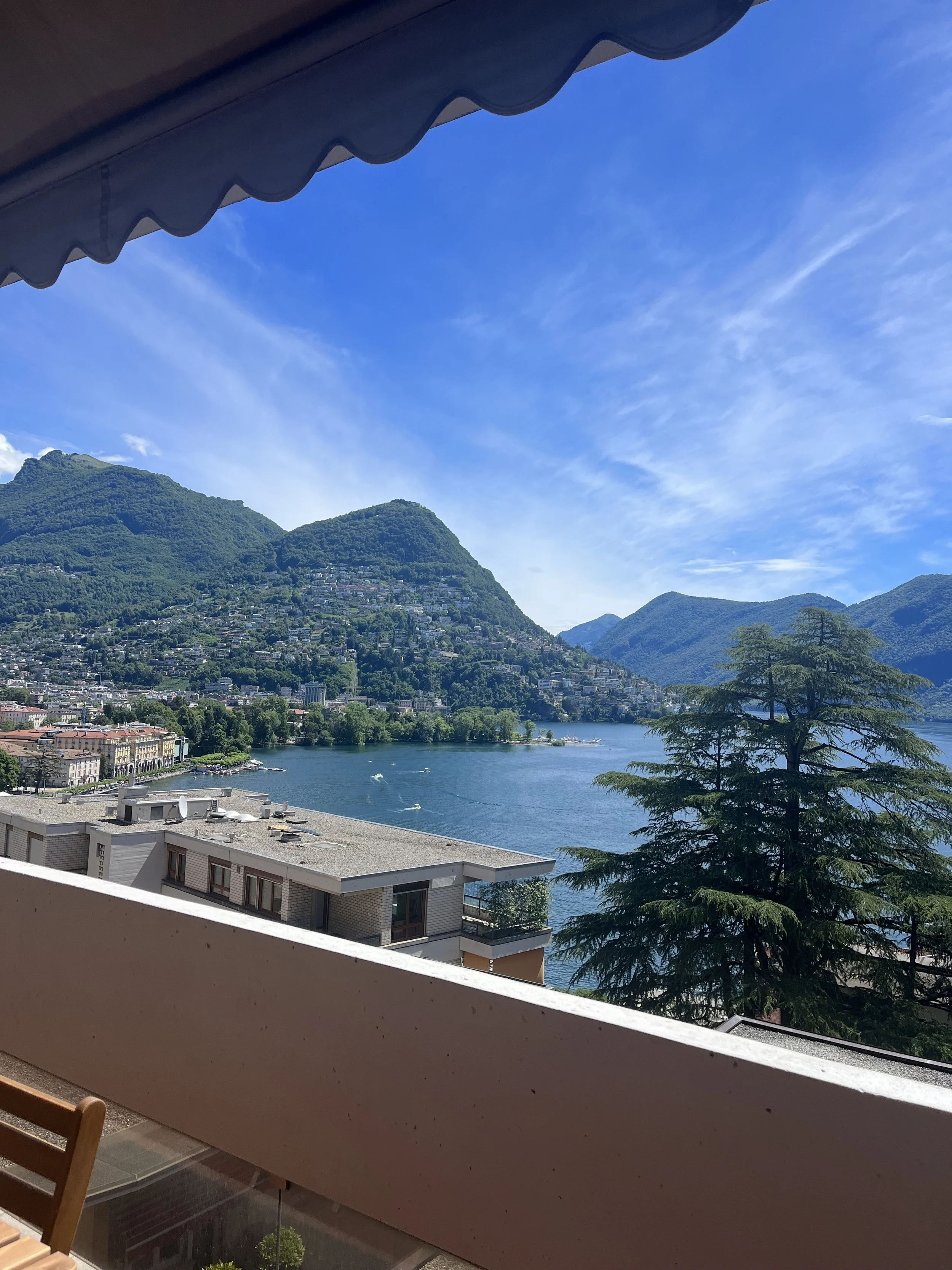 View from a balcony showing a lake surrounded by green hills and mountains under a blue sky with wispy clouds, partially covered by a scalloped awning.