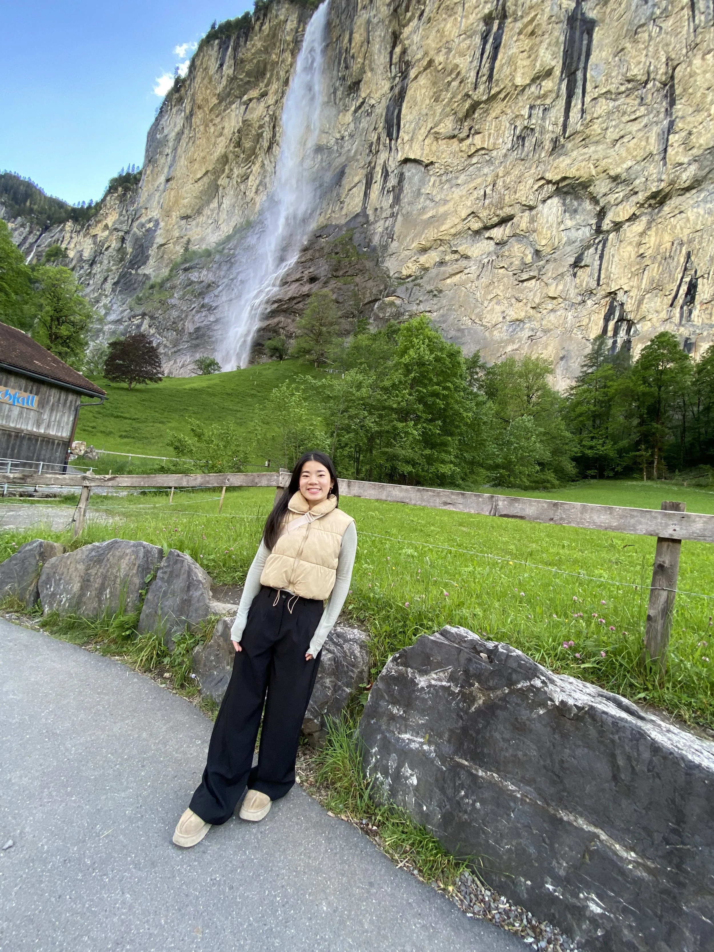 A smiling woman standing in front of a rocky hillside with a tall waterfall cascading down the cliffside. There are trees and a grassy area behind her, and a wooden fence with large rocks at the base along the paved pathway.