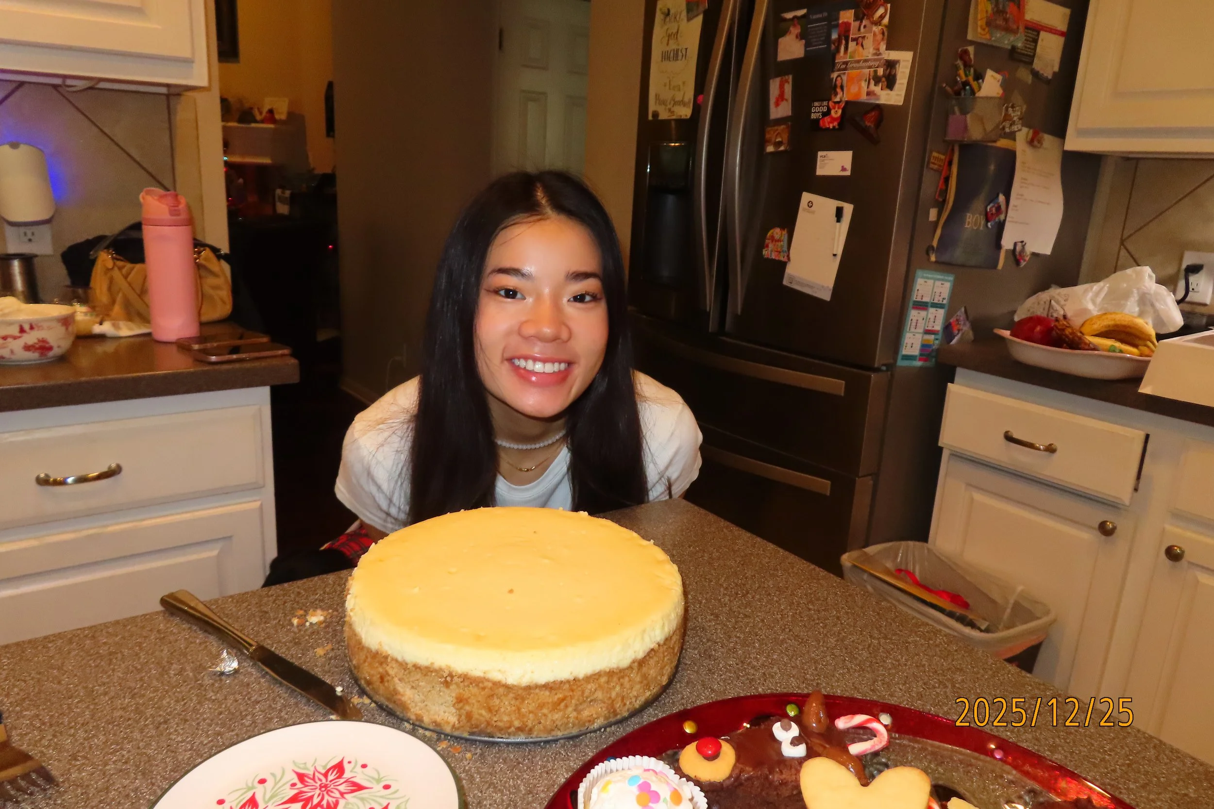 A woman with long black hair smiling at a kitchen counter with a large round cheesecake in front of her, decorated for a celebration. There are various decorated cookies and candies on the counter. The kitchen has white cabinets and a stainless steel refrigerator covered with lots of photos, notes, and magnets. The woman is wearing a white shirt and a pearl necklace.