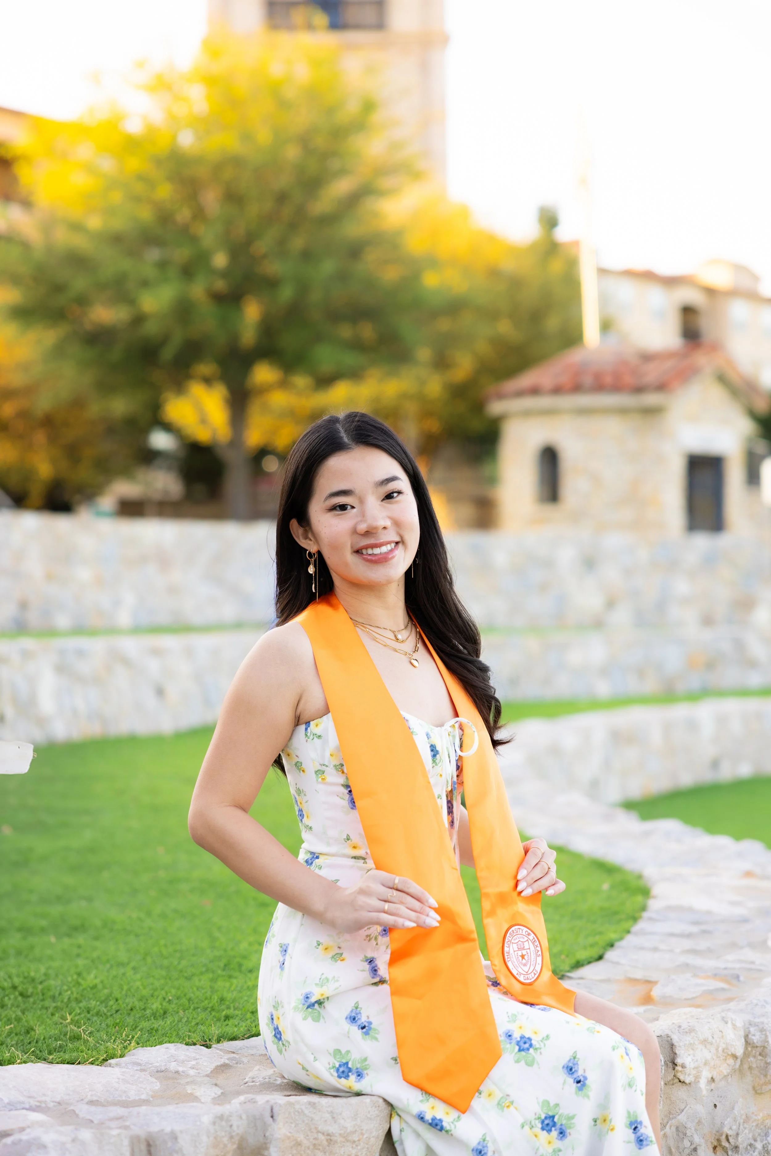 A young woman in a white floral dress with blue and yellow flowers, sitting on a stone ledge outdoors. She is wearing an orange sash and a matching orange honor cord, with a university emblem. She has dark hair, gold earrings, and layered necklaces, and is smiling at the camera. The background features a stone wall, green grass, and trees with yellow leaves, with a building in the distance.