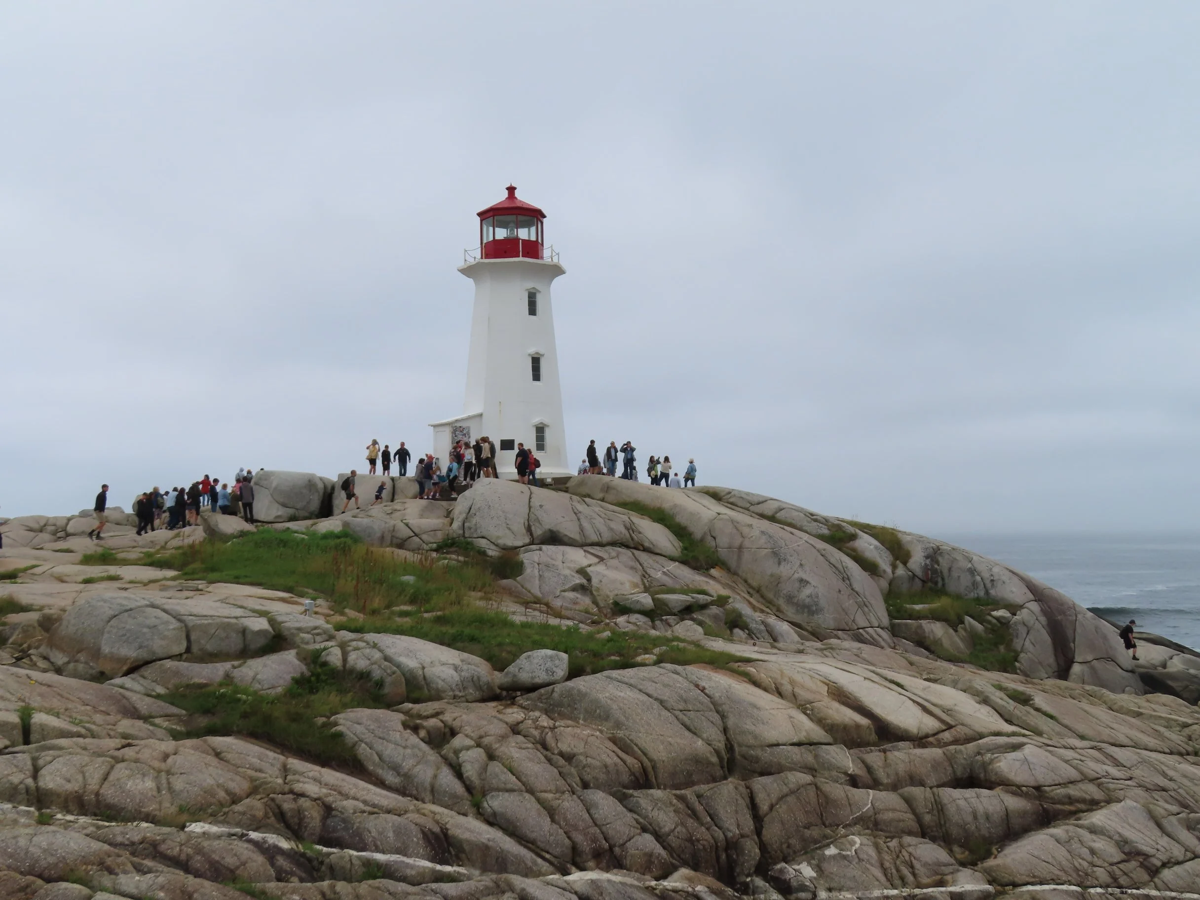 A white lighthouse with a red top stands on rocky terrain by the ocean, with a crowd of people gathered around and visiting the site.