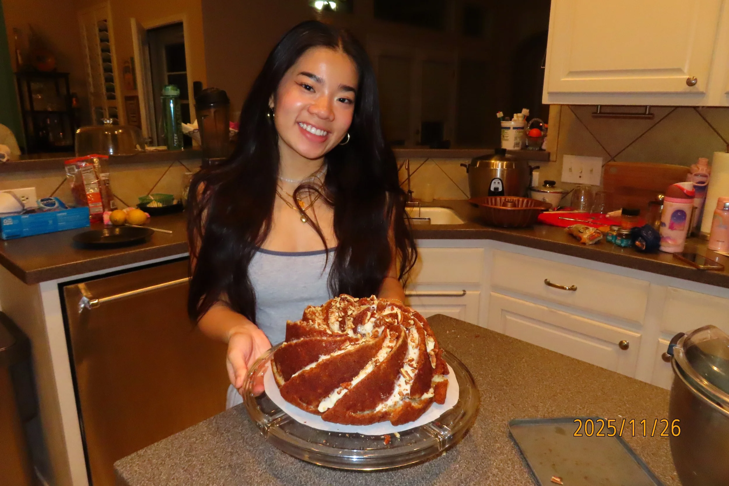 Smiling woman in a kitchen holding a plate of a large homemade cake with chocolate and cream frosting.