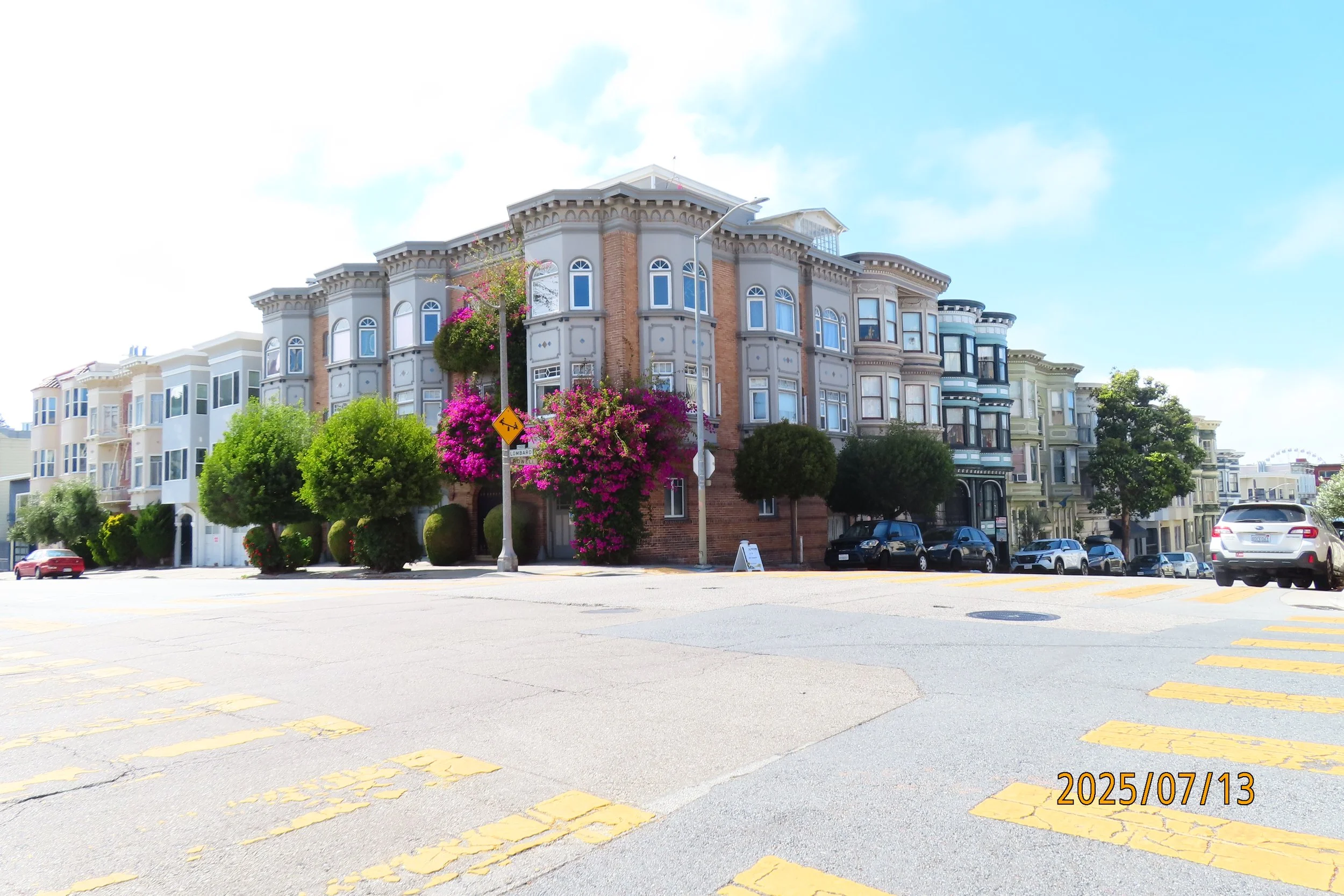 A street scene with a row of colorful Victorian-style houses, trees, parked cars, and a bright blue sky with some clouds.