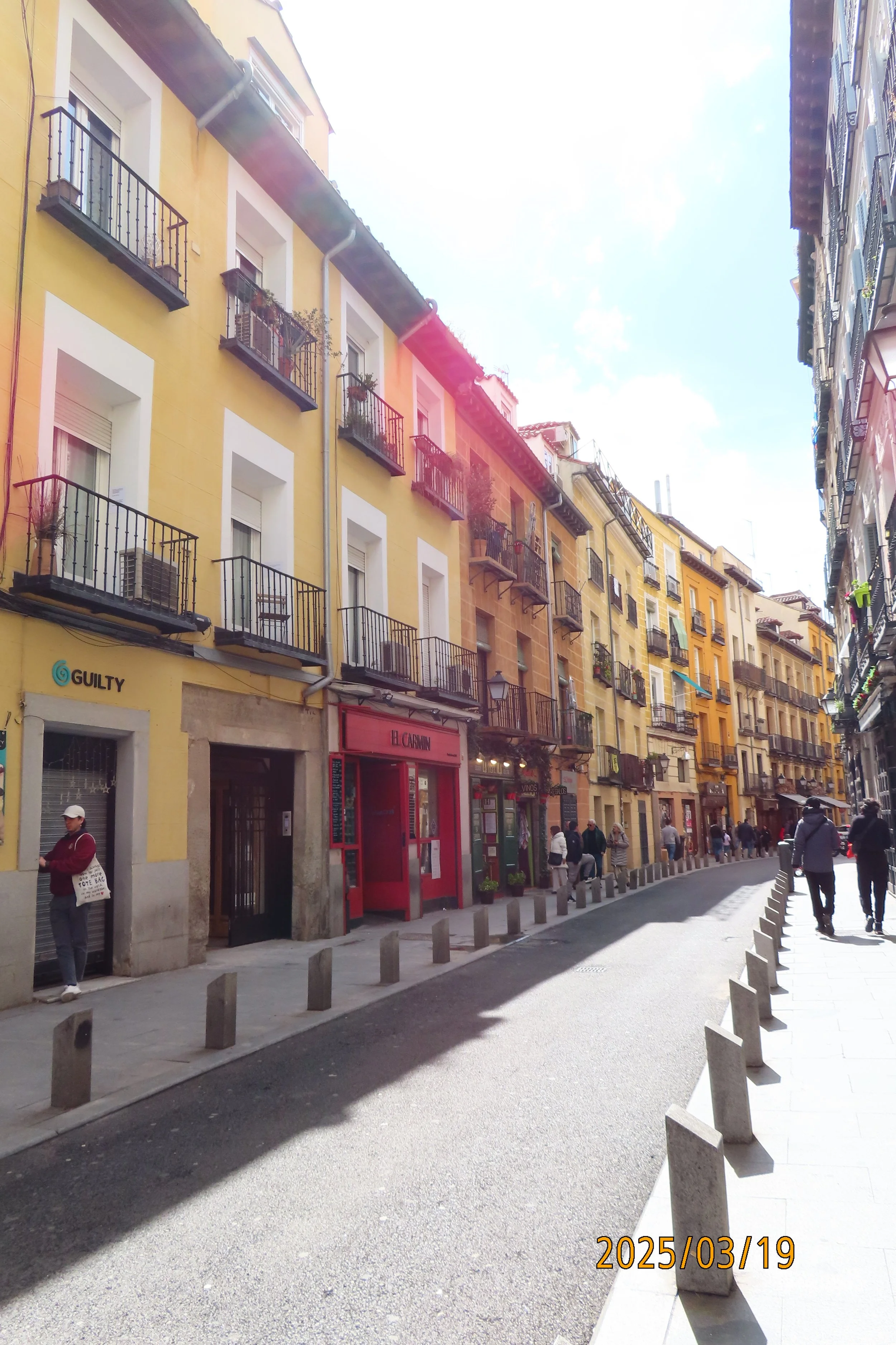 Street view with colorful buildings featuring balconies and shops, pedestrians walking, and sunlight casting shadows.