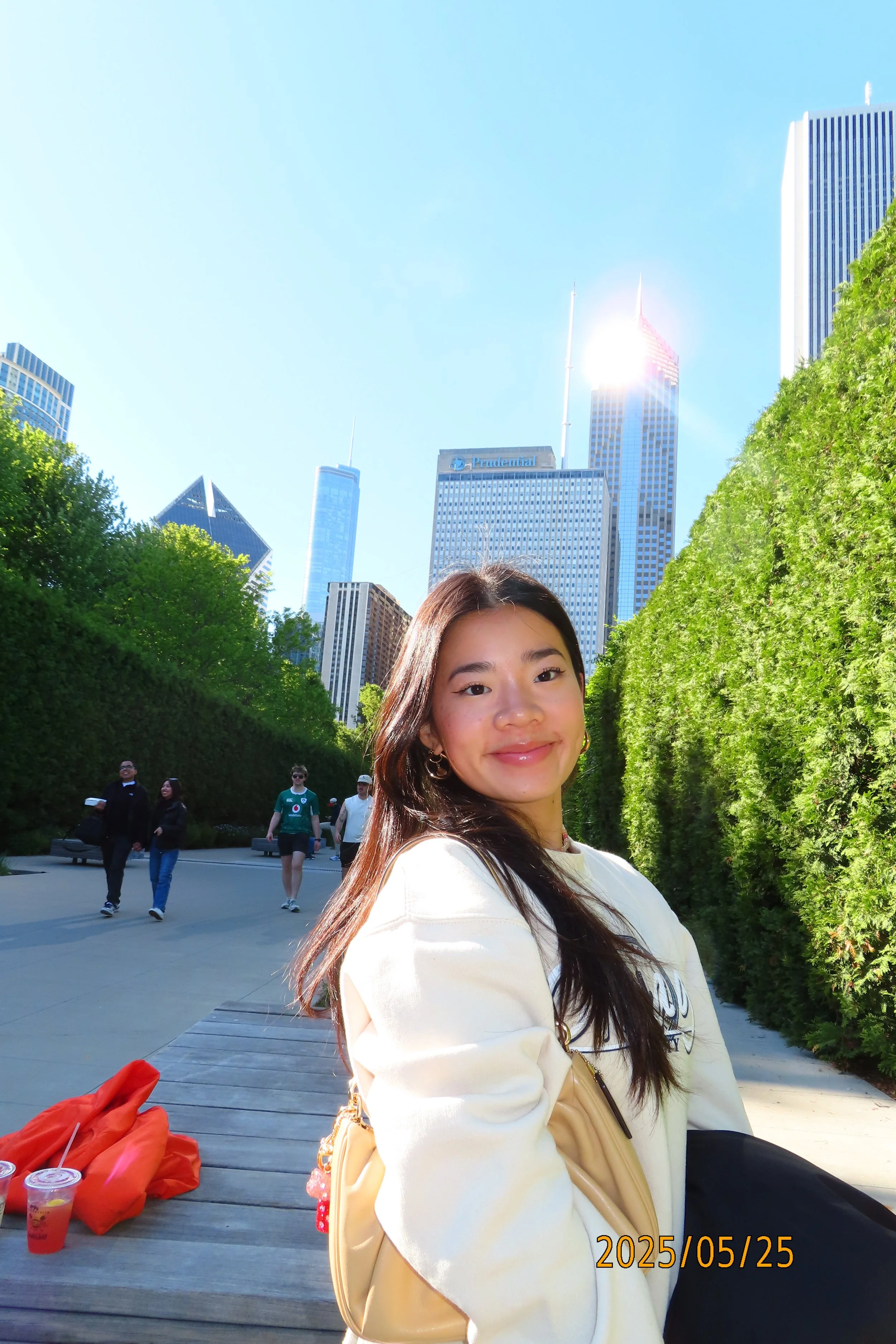 A young woman smiling outdoors with a city skyline and tall buildings in the background on a sunny day, with a date stamp of May 25, 2025.