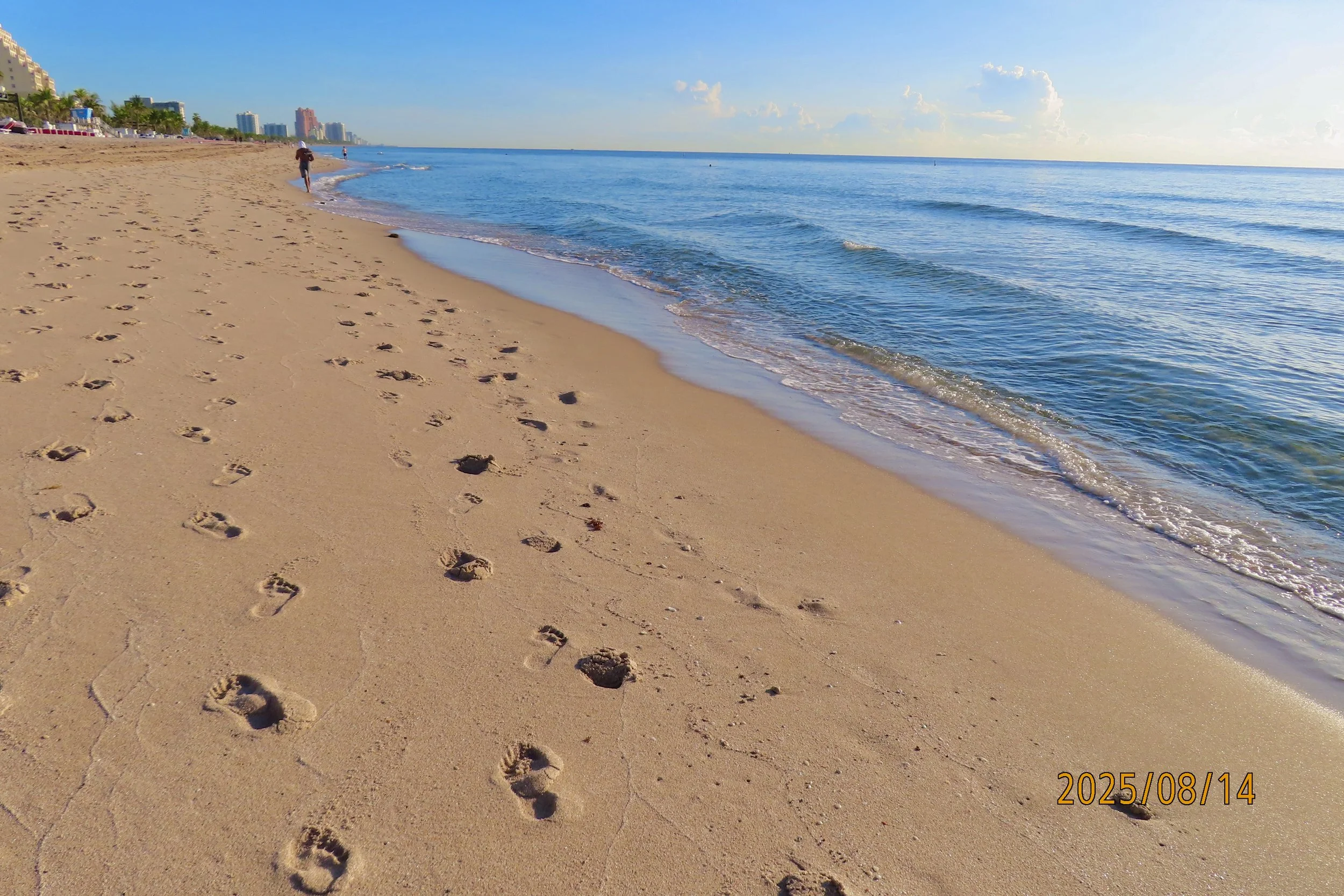 Footprints in the sand on a beach, with calm ocean waves and a clear blue sky in the background, along with some high-rise buildings in the distance.