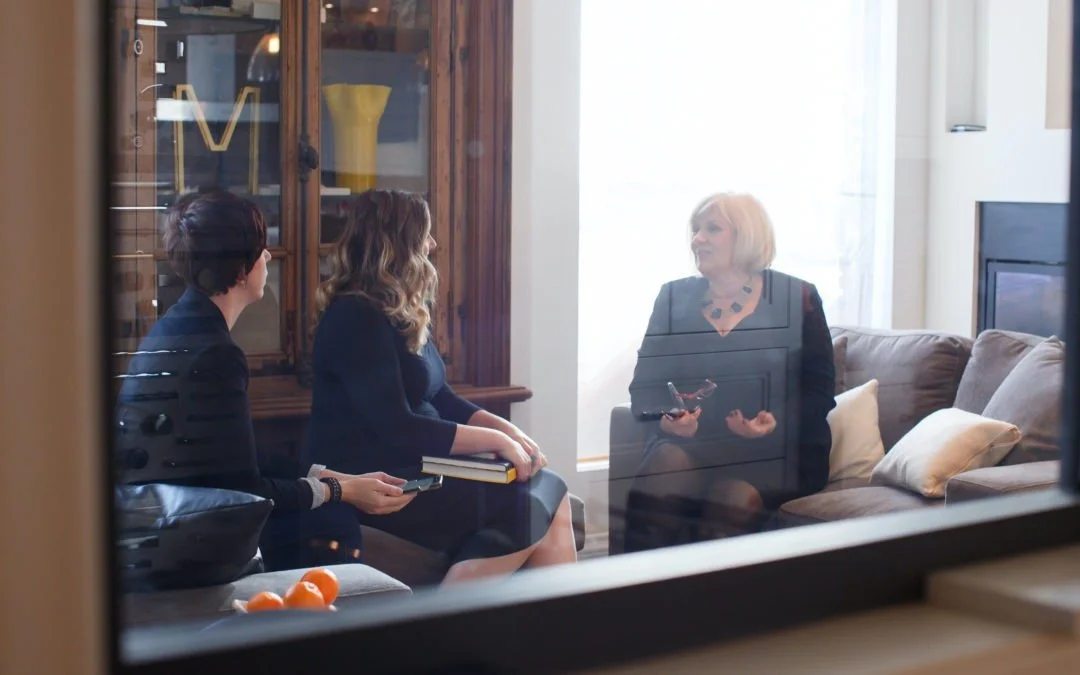 Three women having a conversation in a living room, viewed through a window. One woman is sitting on a sofa with a notepad, another is sitting on a chair holding a notebook, and the third woman is standing or sitting on the floor, speaking.