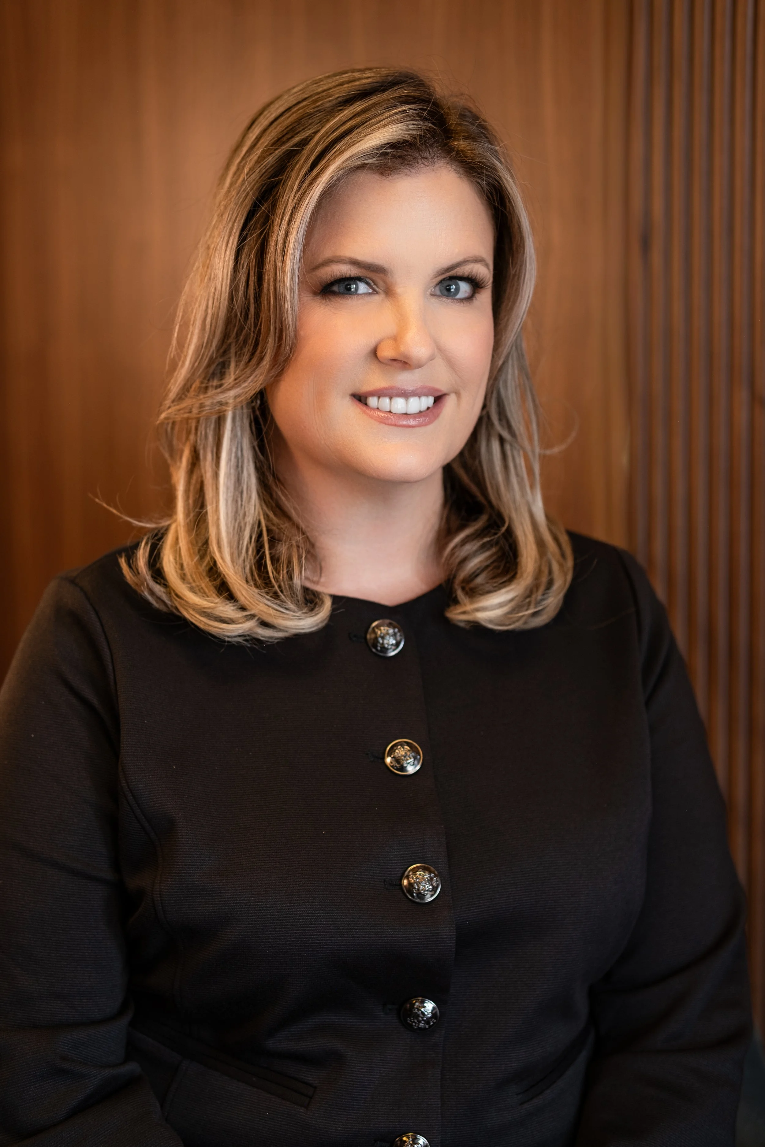 A woman with blonde hair and blue eyes, wearing a black blazer with decorative buttons, sitting indoors with a wooden panel background.