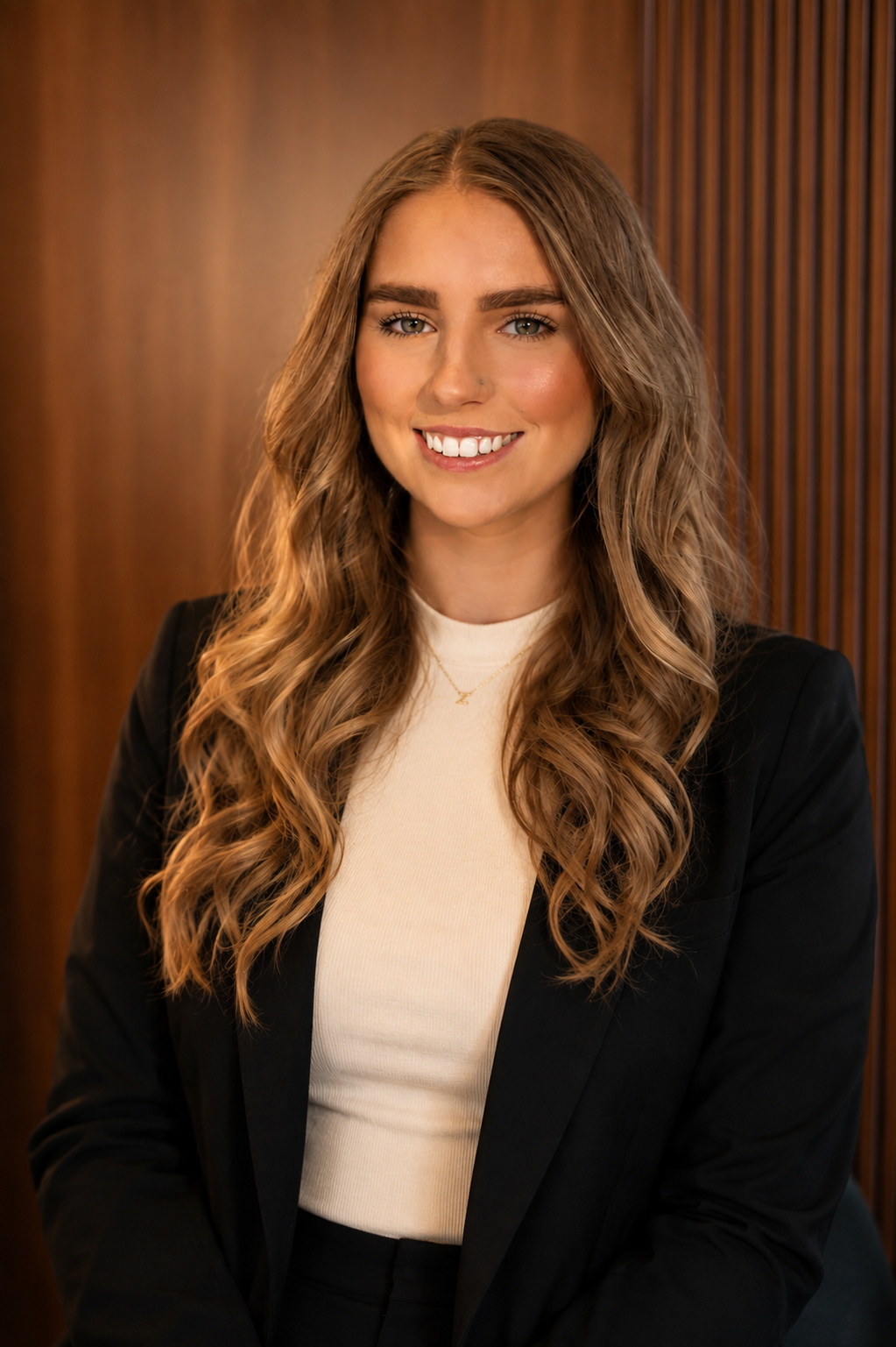A young woman with long, wavy blonde hair and a nose piercing, wearing a black blazer and a cream turtleneck, smiling in front of a wooden background.