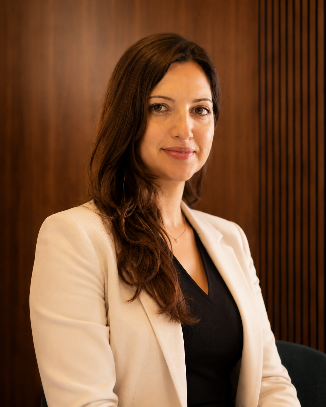 Businesswoman with brown hair, wearing a beige blazer and black top, sitting in an office with wooden paneling.