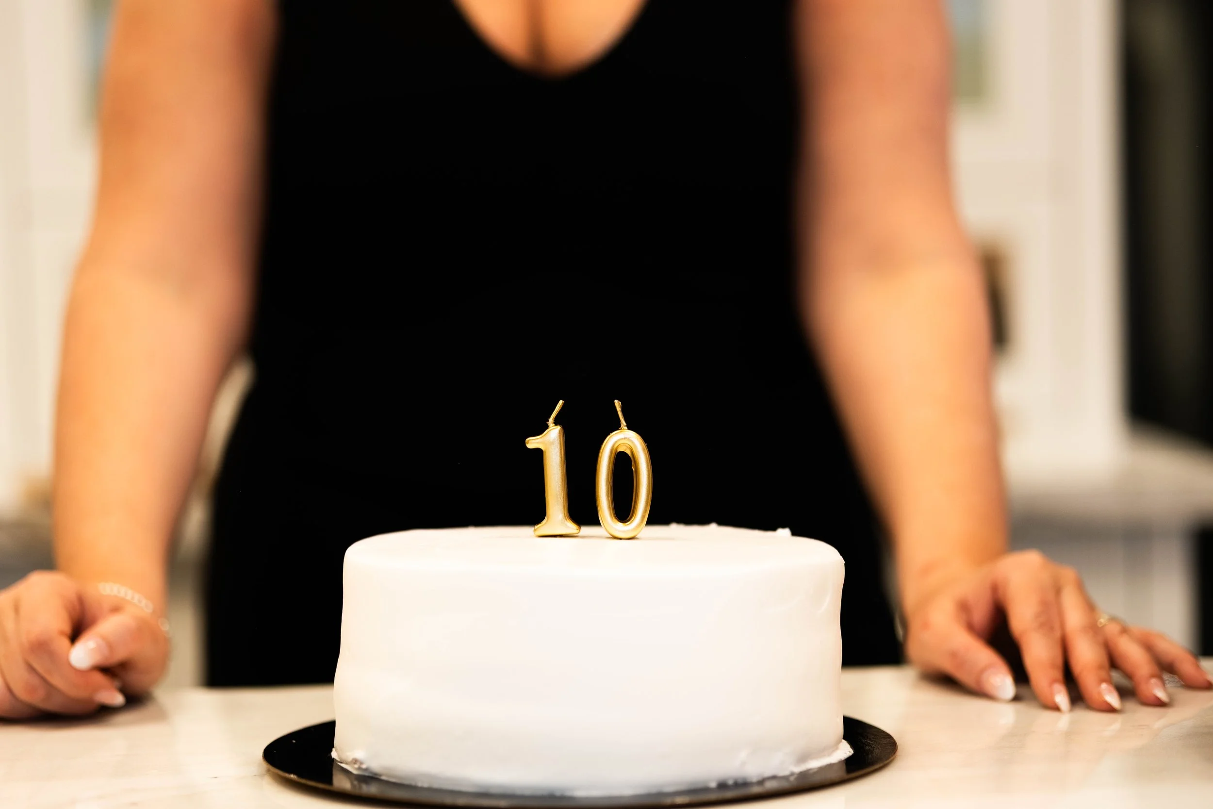 Person in a black dress holding a white cake with gold number candles 10 on top, celebrating a 10th birthday.