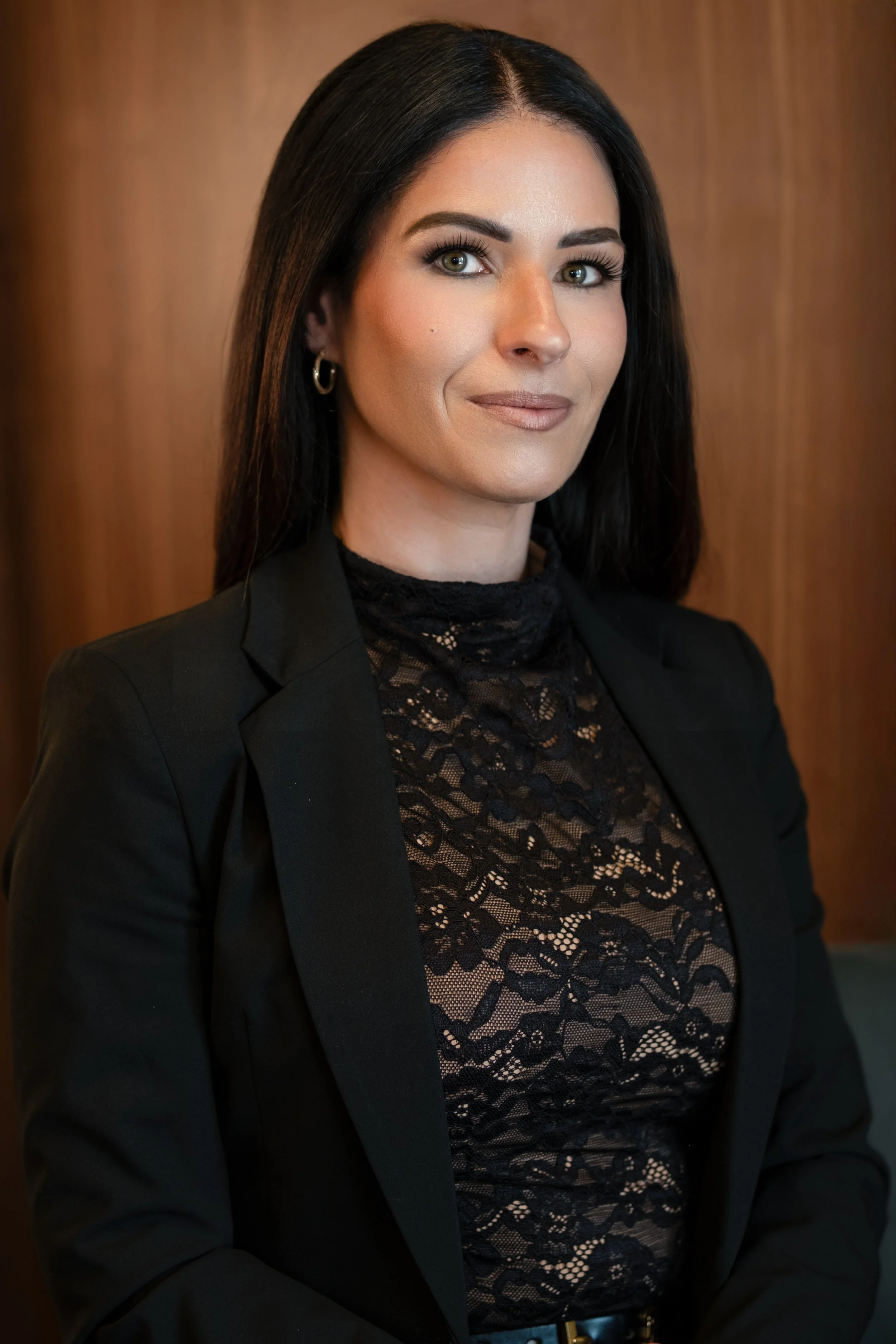 A woman with long black hair, wearing a black blazer over a black lace top, posing in front of a wooden background.