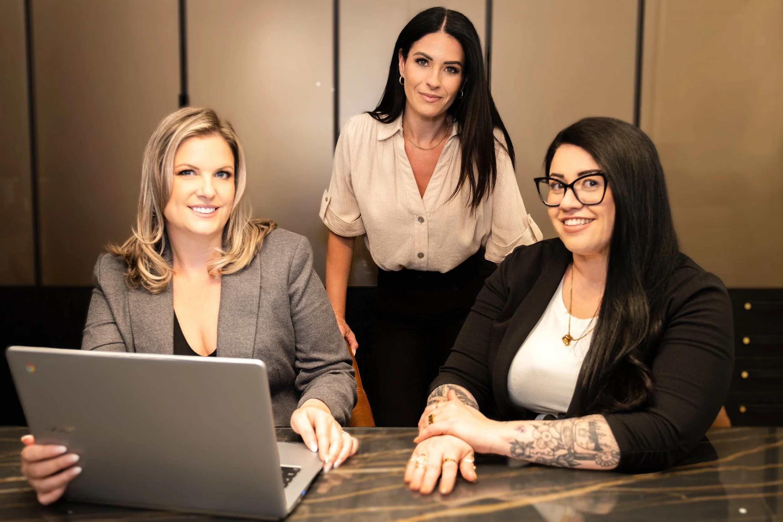 Three women in a business setting gathered around a laptop, with one woman standing and the other two seated. They are smiling and appear engaged in a discussion. The background features a neutral wall.