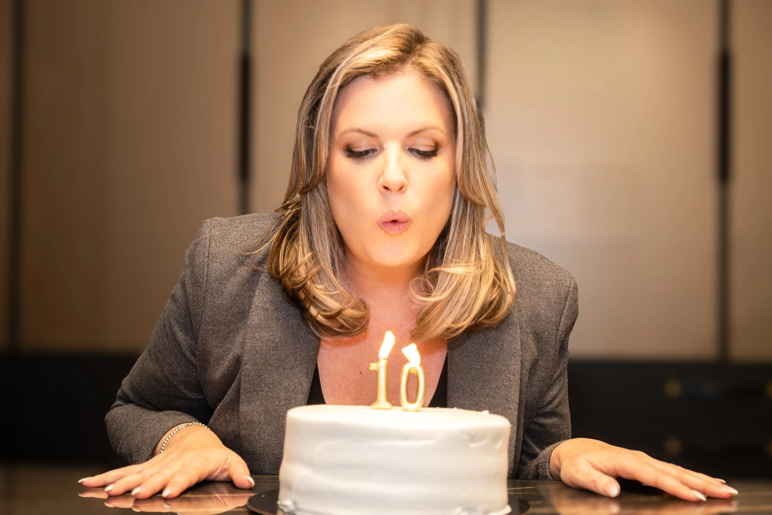 A woman with blonde hair and a gray blazer blowing out candles on a birthday cake with a '10' candle.