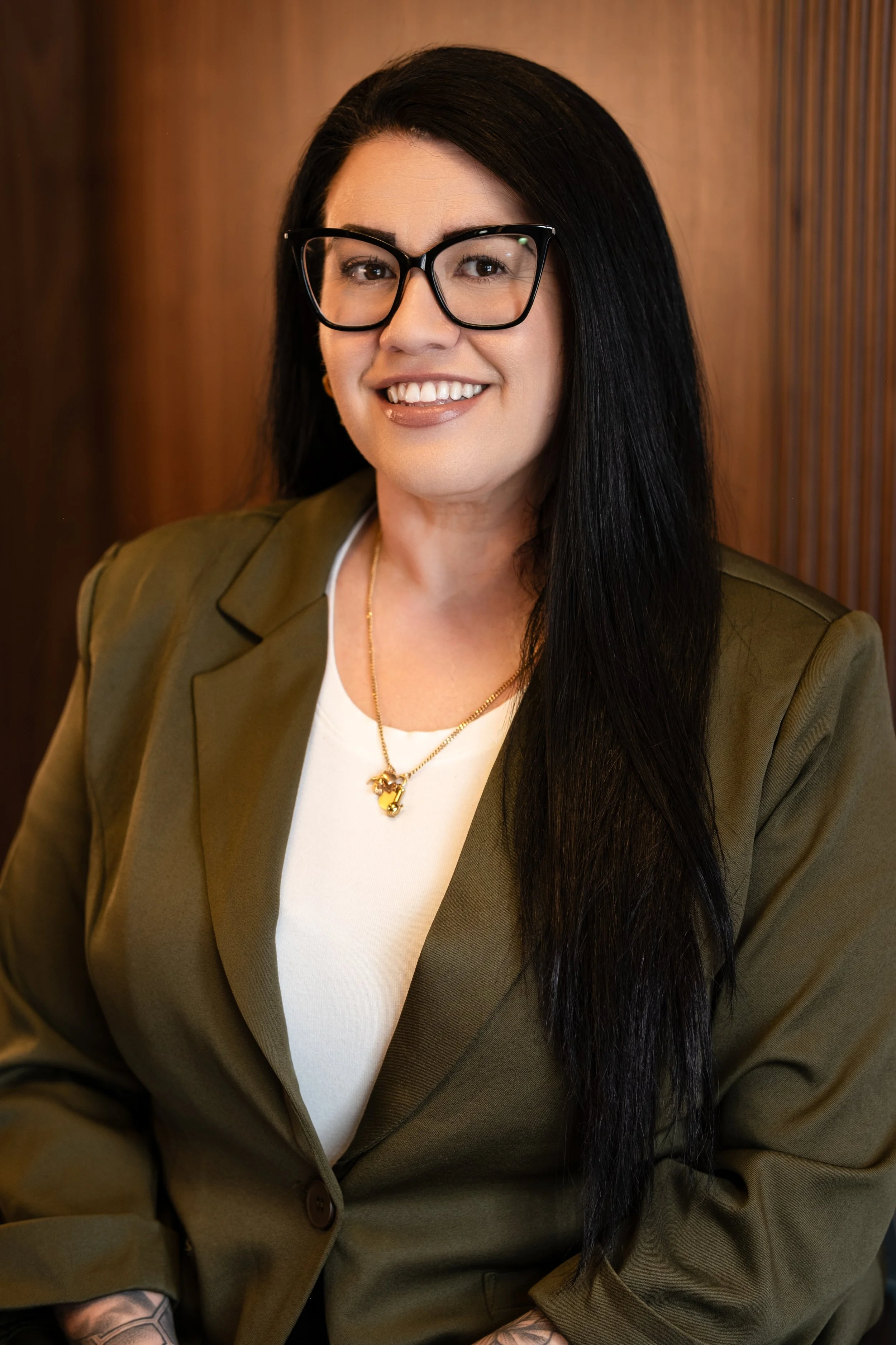 A woman with long black hair, glasses, and a olive green blazer, smiling, sitting against a wooden background.