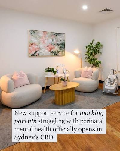 Modern waiting room with two rounded armchairs with pink pillows, a small round wooden table, and a tall plant in the background. Wall art features pink and green floral painting.