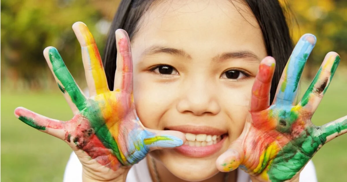 A young girl smiling and holding her hands up, covered in colorful paint, with a blurred outdoor background.