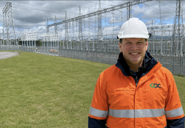 Man in an orange GDC safety jacket and white hard hat standing outside at a power substation with electrical infrastructure and cloudy sky in the background.
