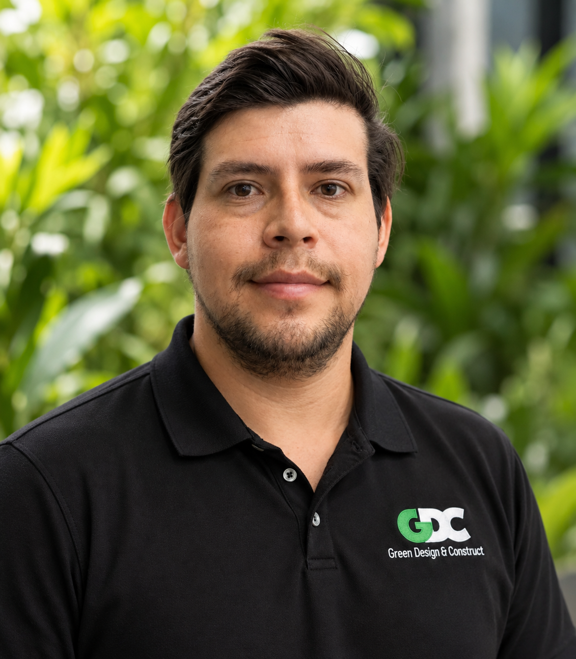 A man with dark hair and a beard, wearing a black polo shirt with the logo 'GDC Green Design & Construct', stands in front of green plants outdoors.