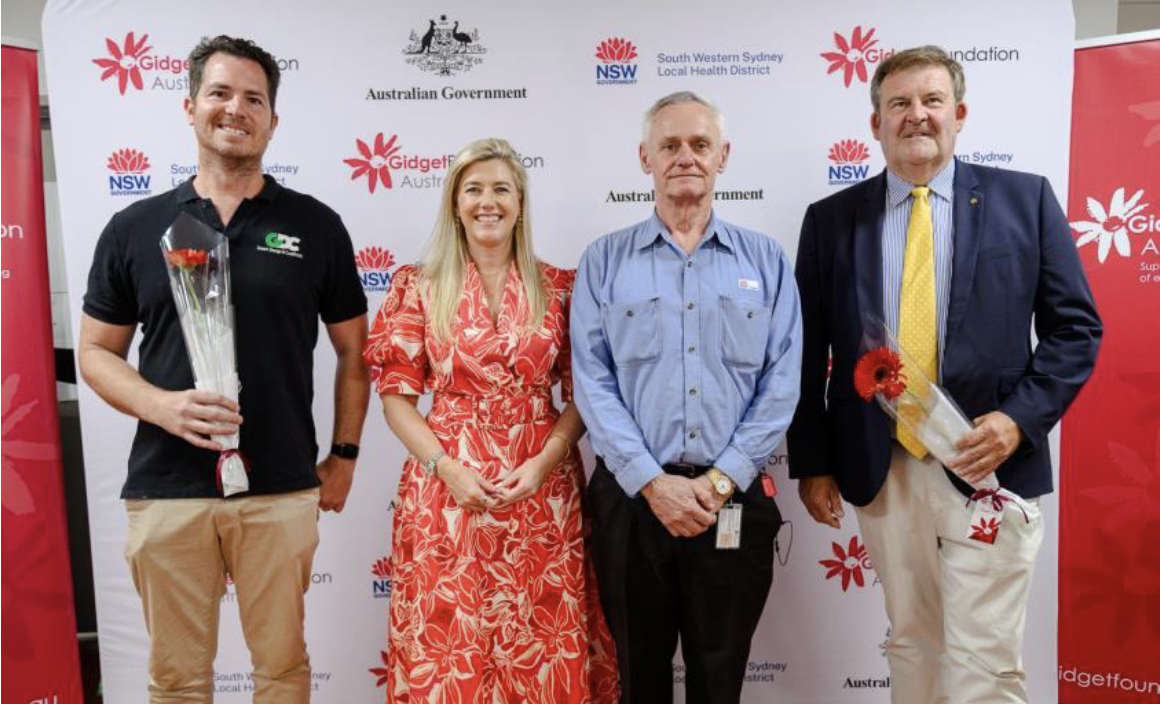 Four people standing in front of a backdrop with logos including Gidget Foundation Australia, Australian Government, and NSW South Western Sydney Local Health District. The two men on the ends are holding flowers, and all are smiling.