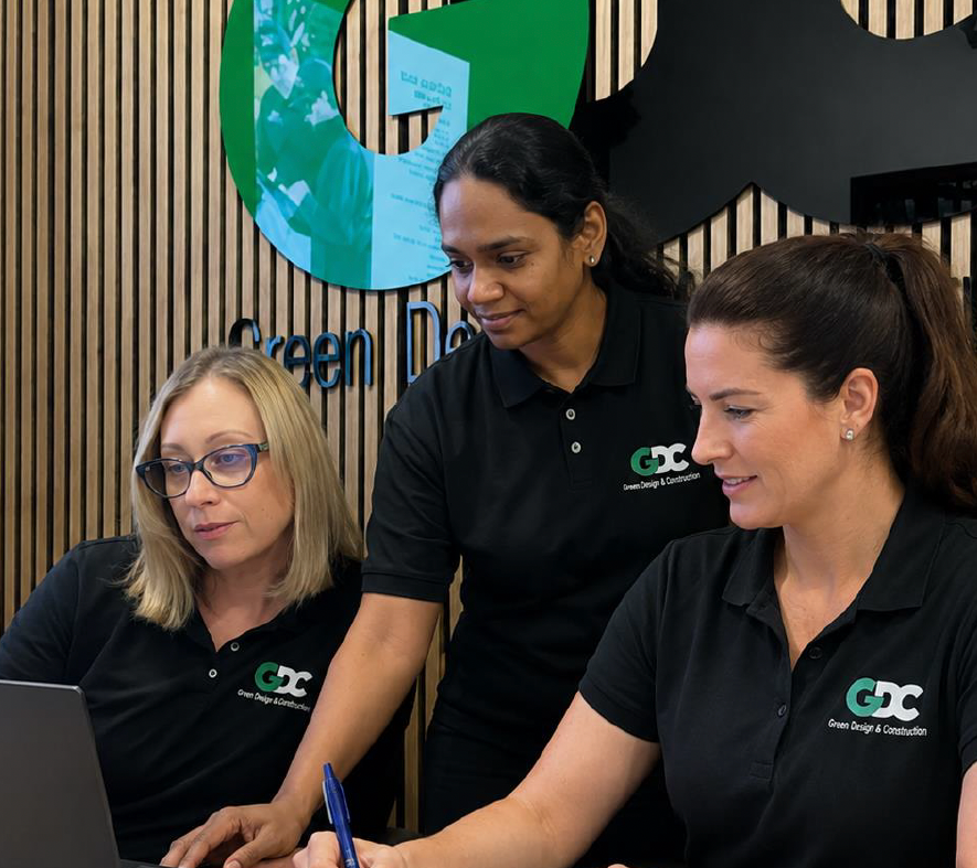 Three women in black GDC shirts working together at a table with a laptop in an office with wood panel walls and a green and blue logo in the background.