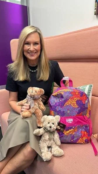 A woman sitting on a pink couch holding two teddy bears, with a colorful patterned backpack and a small pillow beside her.