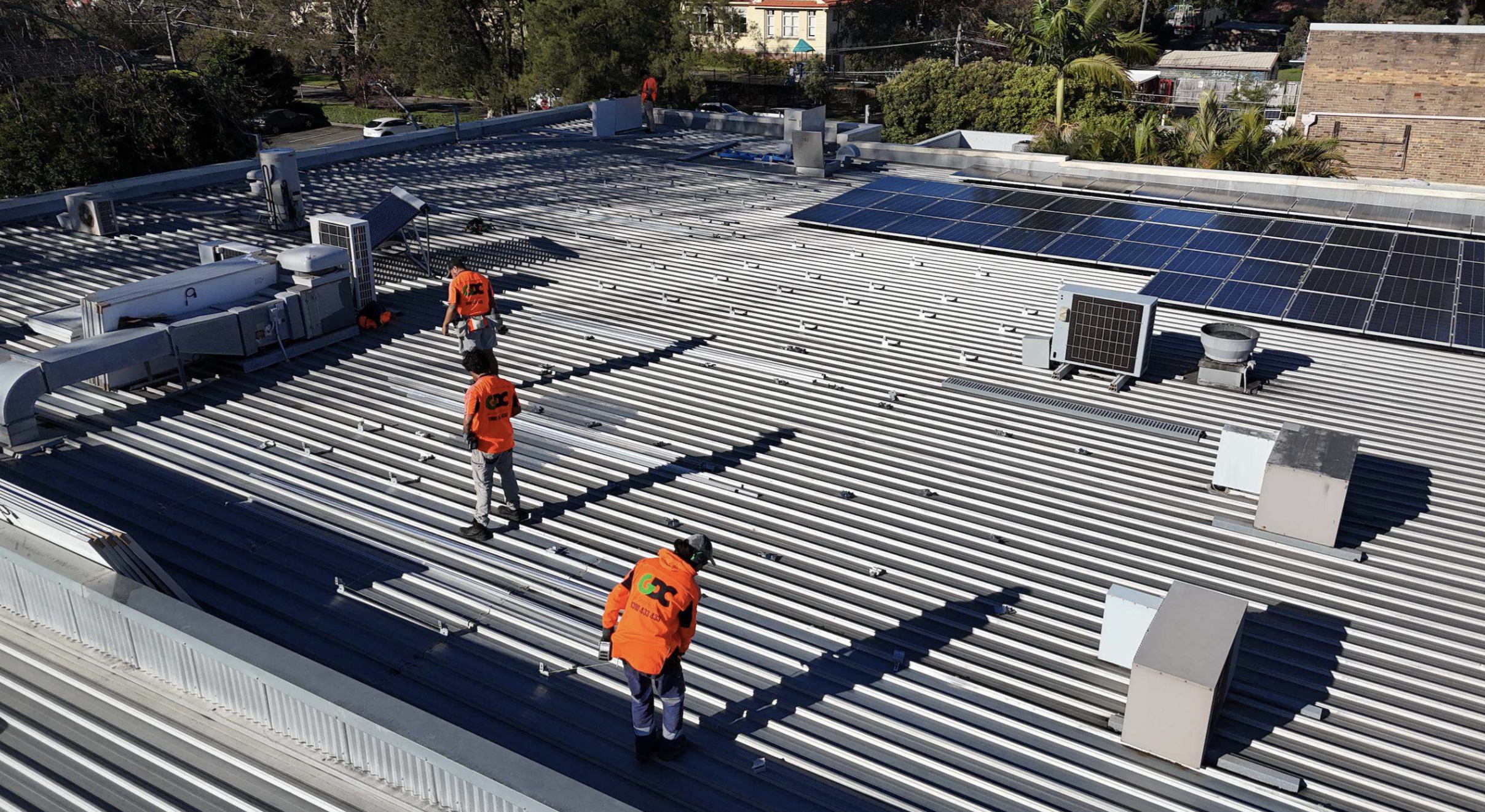 Workers installing solar panels on the roof of a large commercial building with visible HVAC units and metal roofing.