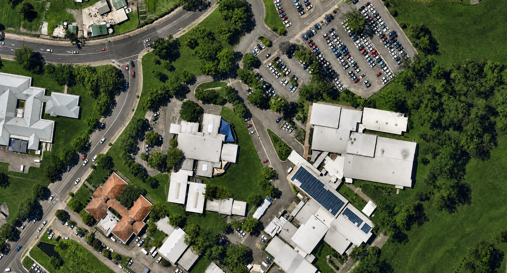 Aerial view of a residential area with multiple buildings, parking lots filled with cars, winding roads, lush greenery, and some buildings with solar panels on the roofs.