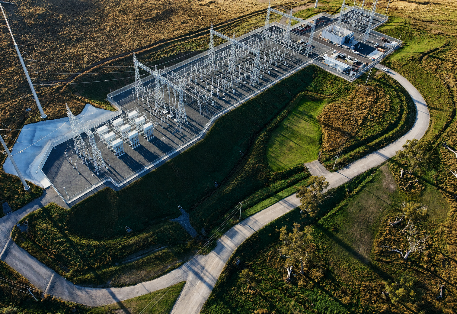 Aerial view of a power substation in a landscape with grassy hills, power lines, and a winding road.