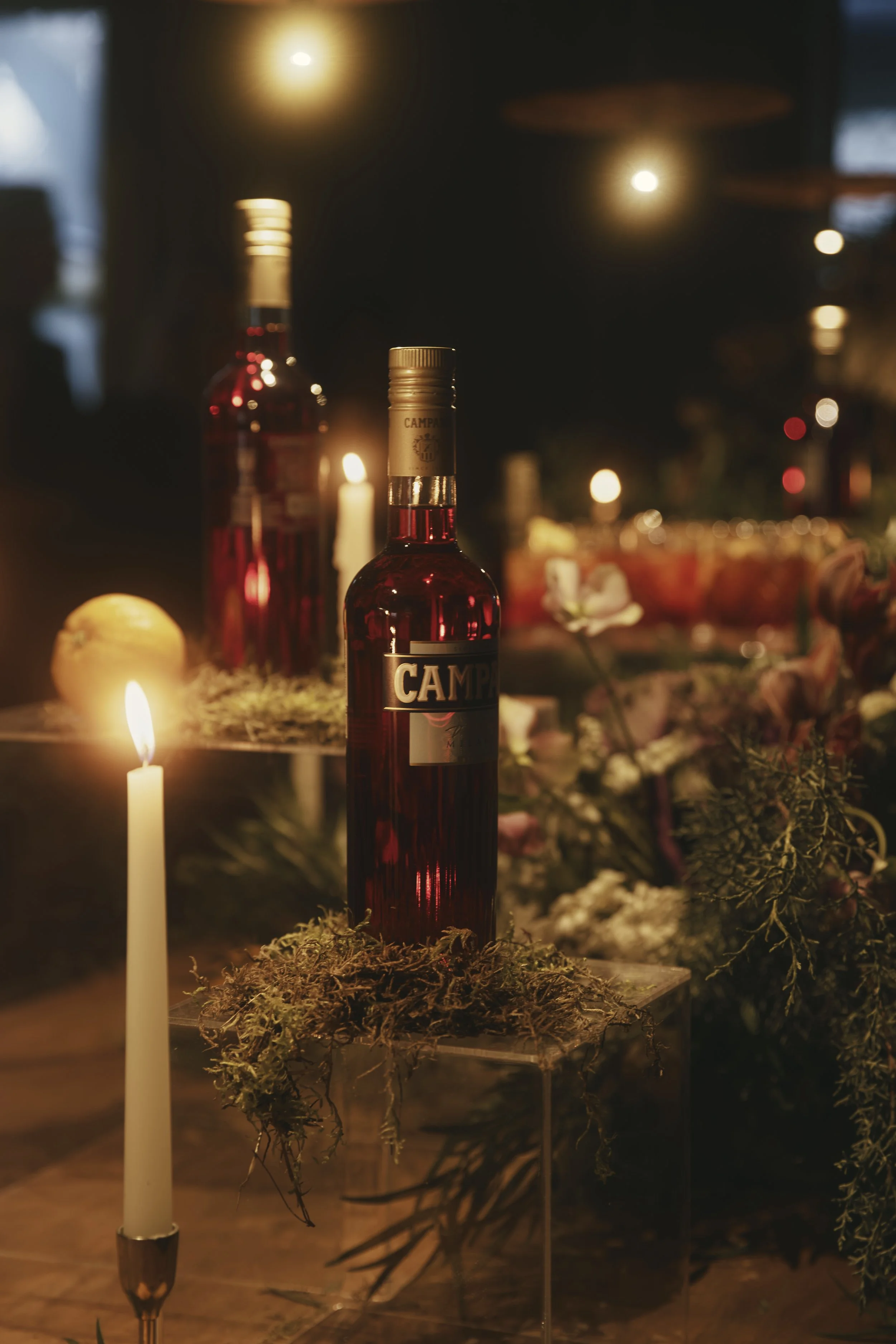 A close-up of a bottle of Campari with candles and greenery around it in a cozy, dimly lit setting.