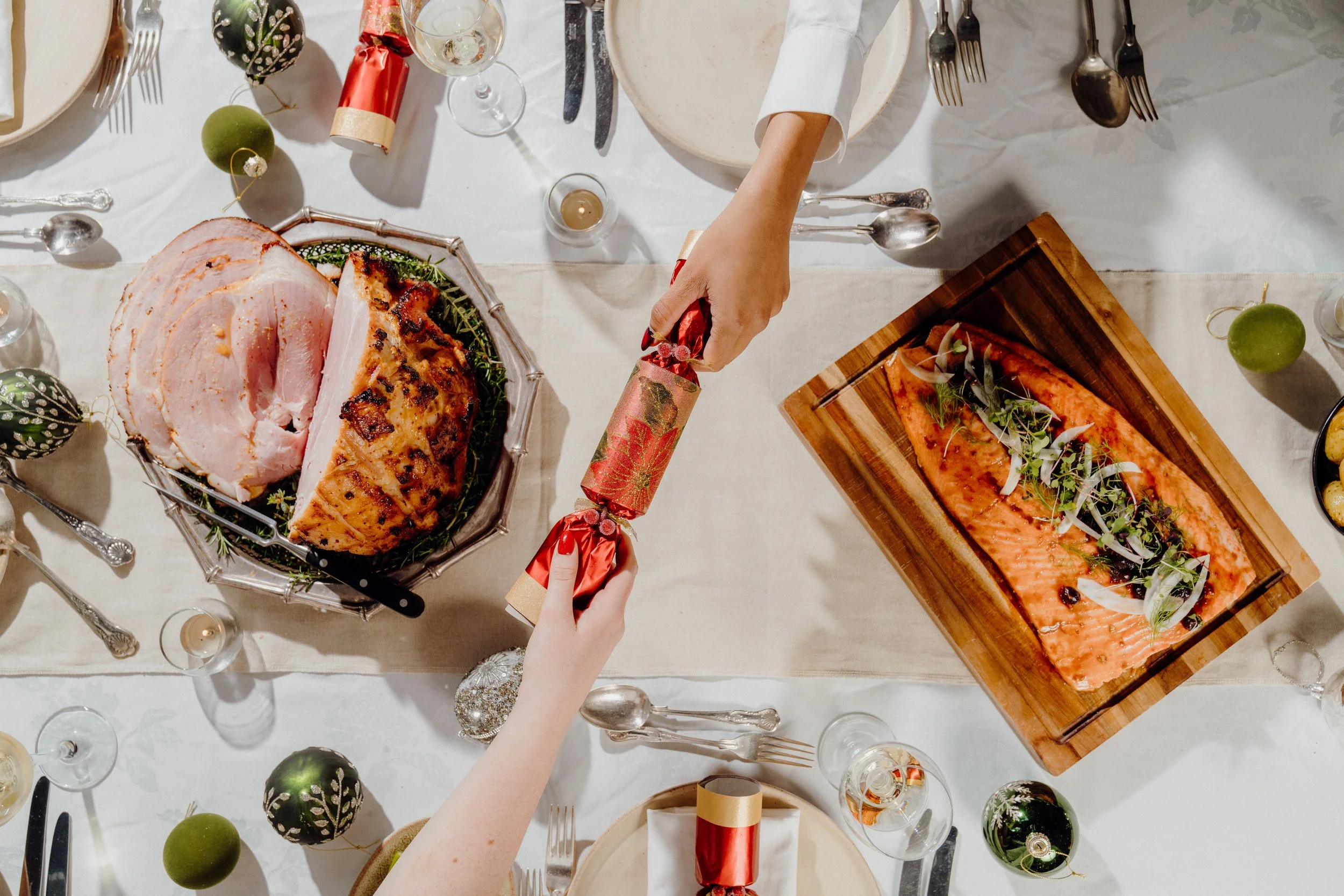 Overhead view of a dinner table with a Christmas theme, featuring a large glazed ham and a baked salmon fillet garnished with herbs. The table is decorated with Christmas ornaments, wine glasses, and candle holders. Two people are in the process of exchanging Christmas crackers.