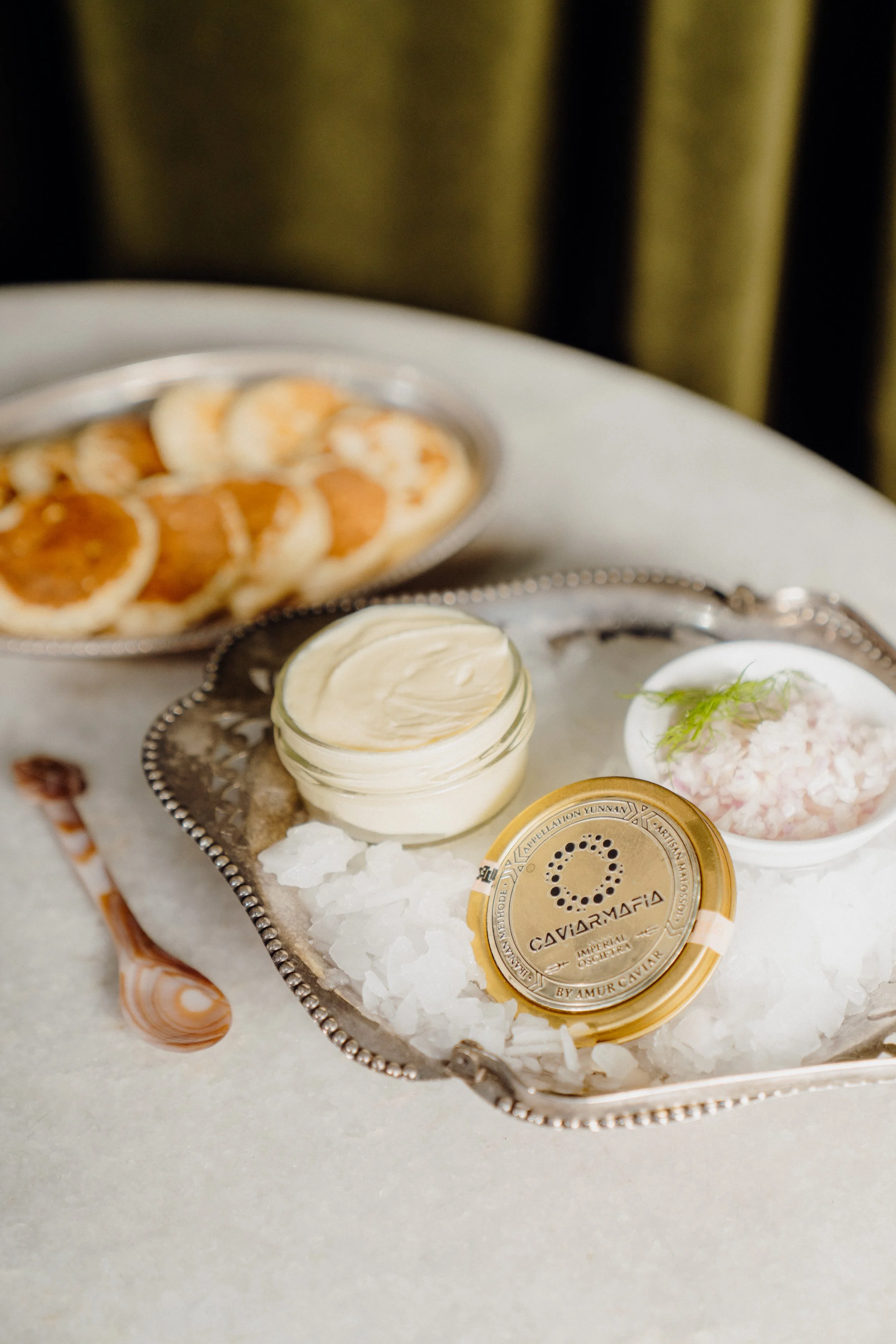 A silver tray with a pearl-like edge holds a small jar of cream, a container of caviar, and a small dish with pinkish spread and greenery, all placed on a bed of white crushed ice.