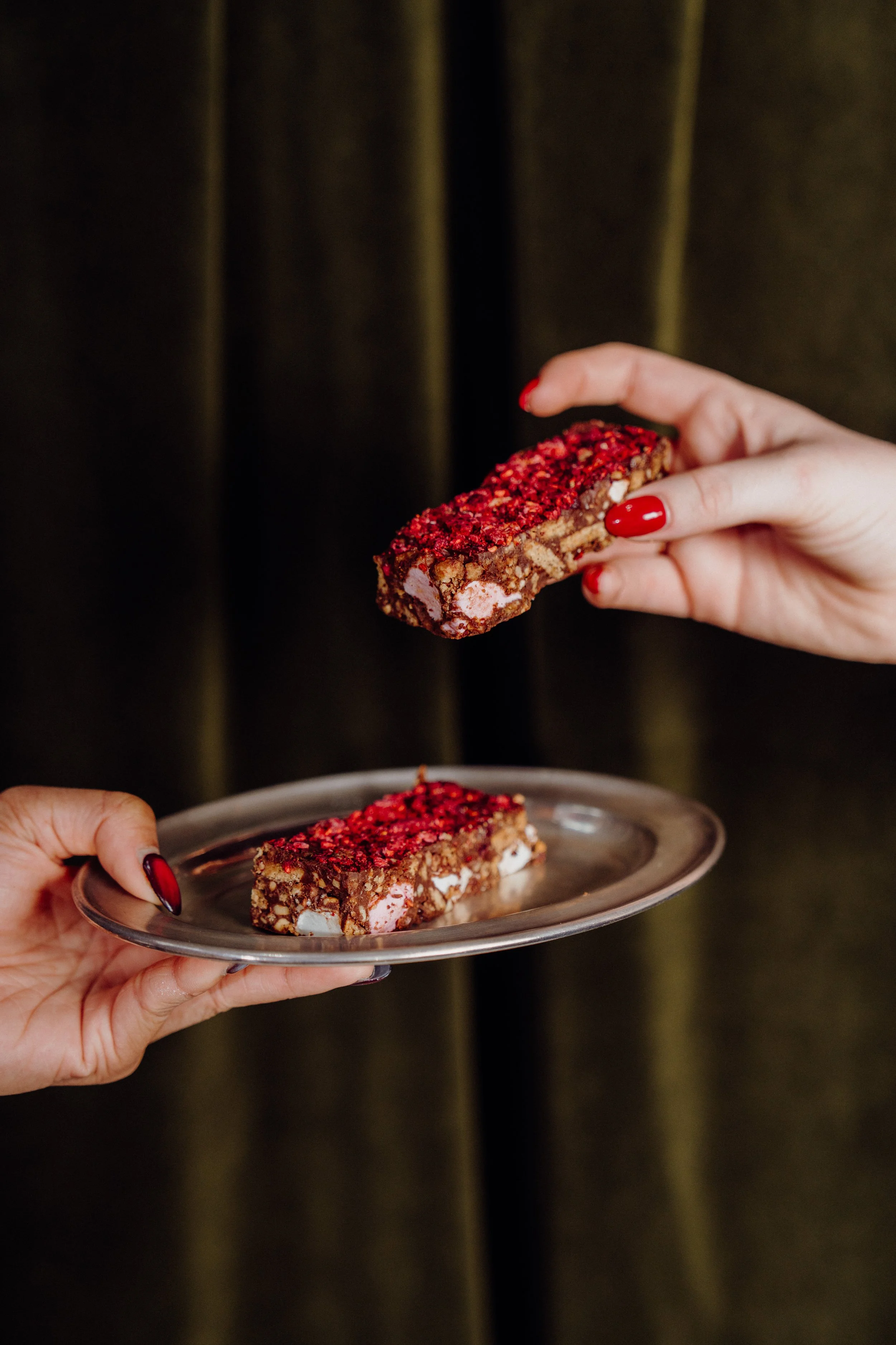 Hands holding a piece of a layered dessert bar with red topping and white pieces, over a silver tray with more bars in the background, against a dark curtain background.