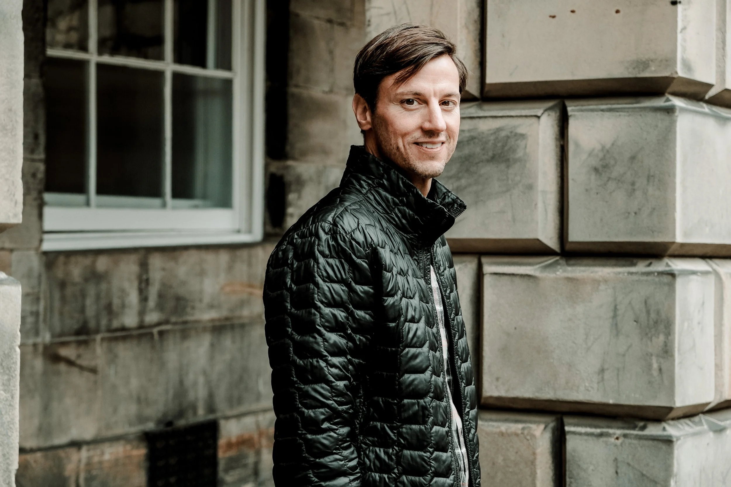A man with brown hair and wearing a black quilted jacket standing outdoors next to a stone building with a window. Billy J. Crawford. Crawford Ledger Co.