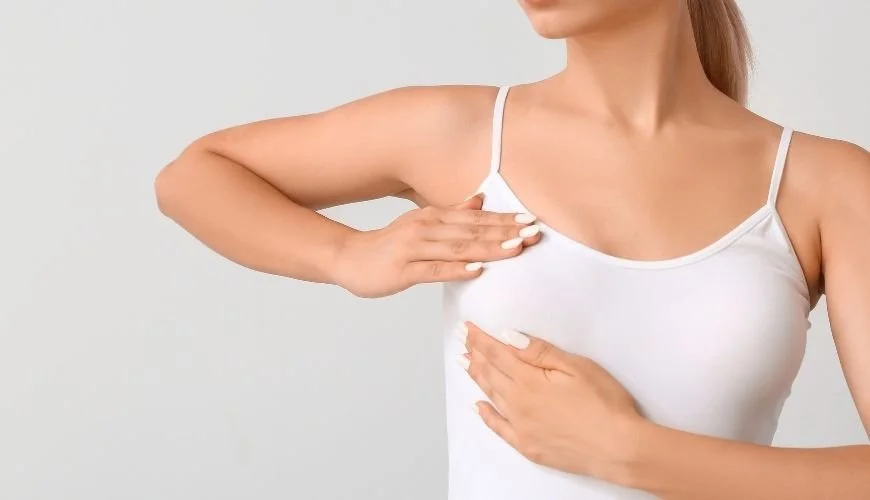 Mujer en camiseta blanca señalando la parte superior del pecho y el lado izquierdo del torso, en un fondo gris claro.