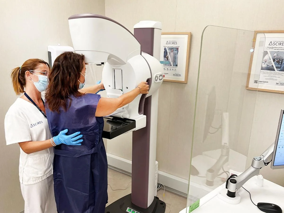 Dos profesionales de la salud operando una máquina de mamografías en una clínica, usando mascarillas y guantes, con paredes de color claro y carteles informativos enmarcados.