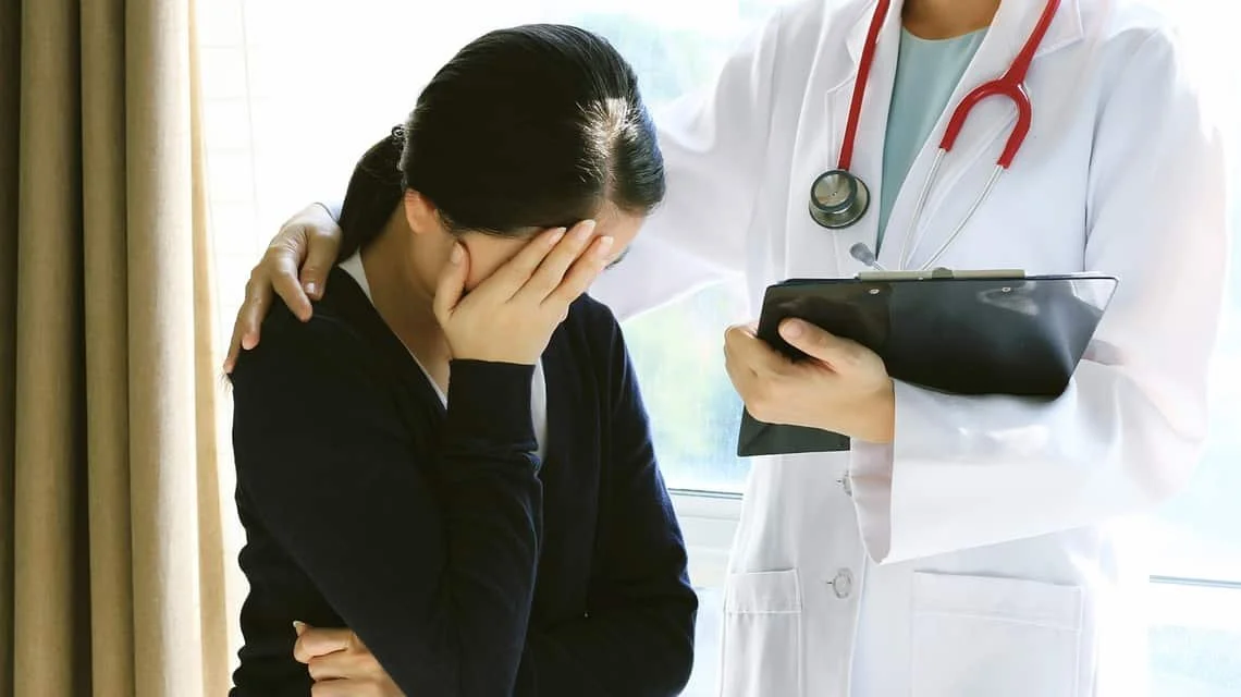 Una mujer con dolor de cabeza siendo atendida por un médico con estetoscopio y un portapapeles.
