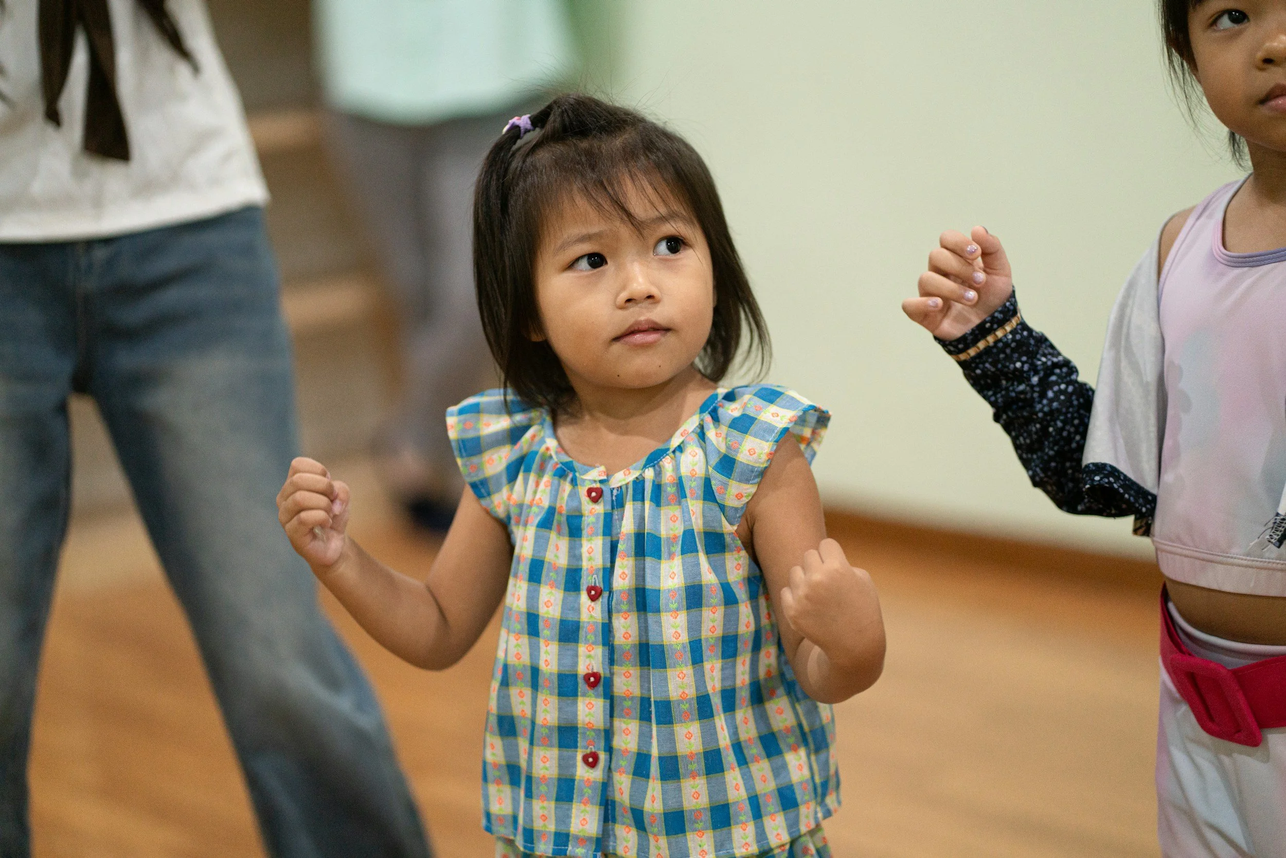 Young girl with short hair and a checkered dress made of blue, yellow, and white colors, with red buttons, raising her fists in a confident pose, participating in an activity with other children in an indoor setting.