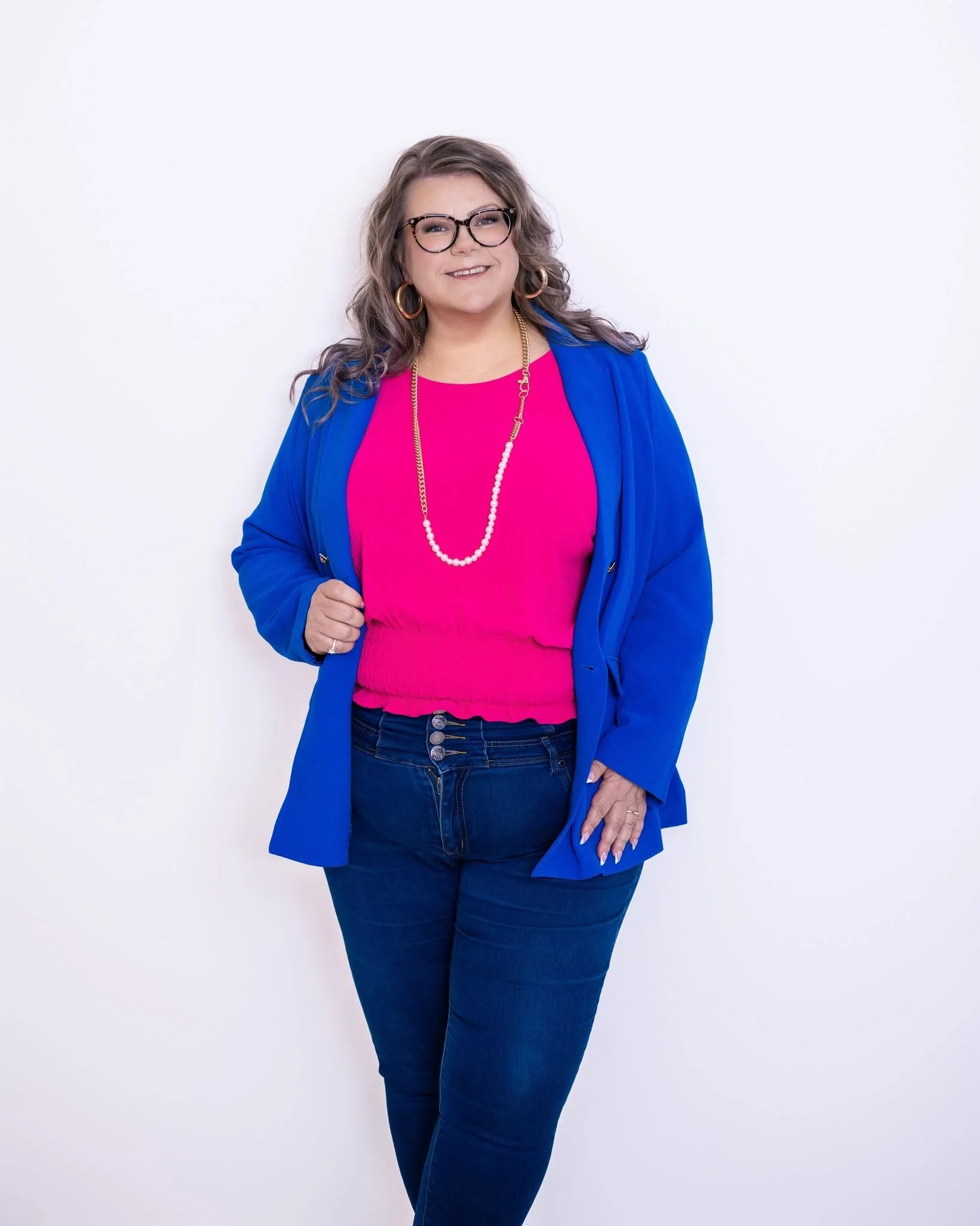 A smiling woman with glasses and large hoop earrings, wearing a bright pink top, a blue blazer, and dark jeans, posing against a white background.