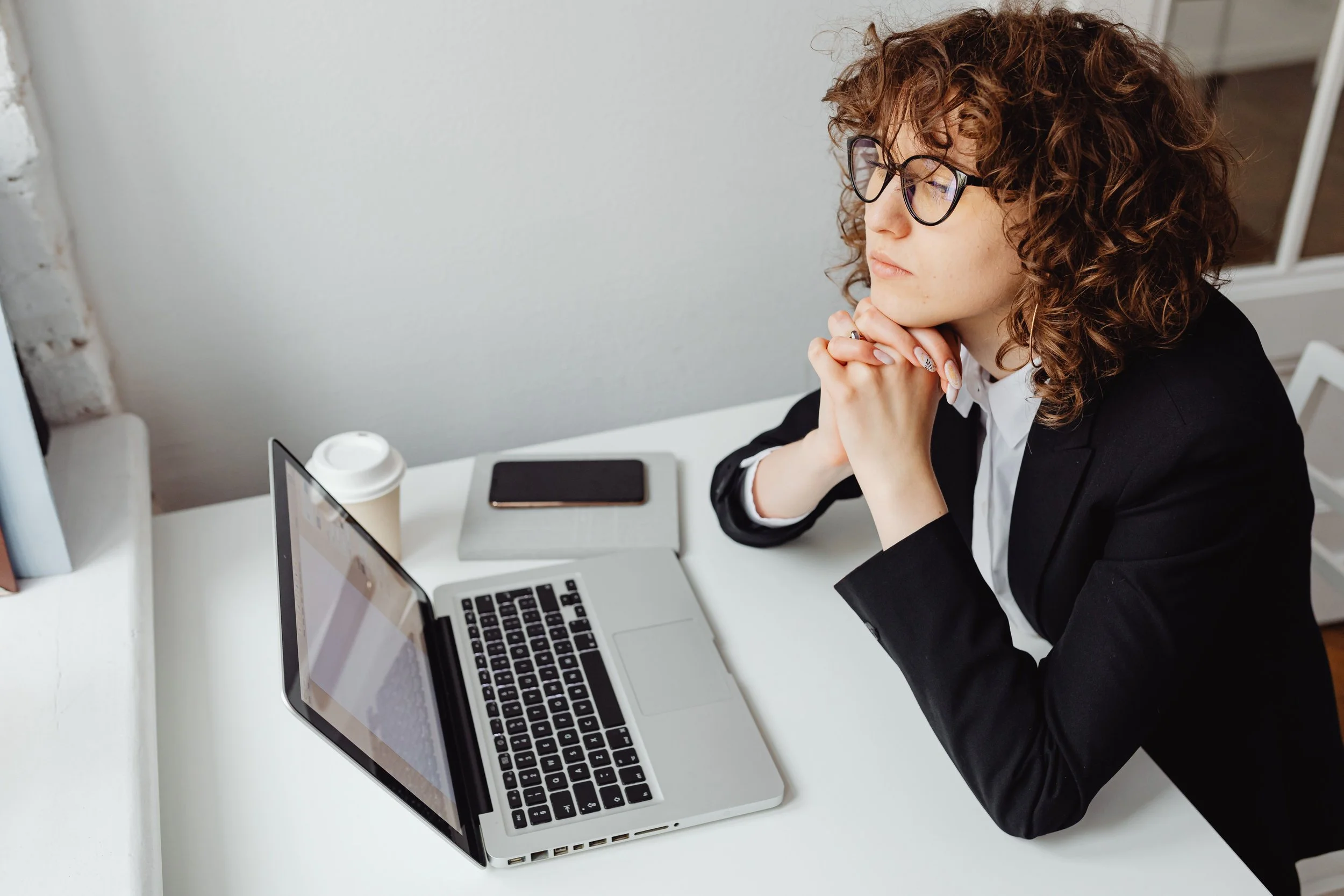 Businesswoman with curly hair and glasses sitting at a white desk, looking at a laptop screen with her hands clasped, beside a disposable coffee cup, tablet, and smartphone.