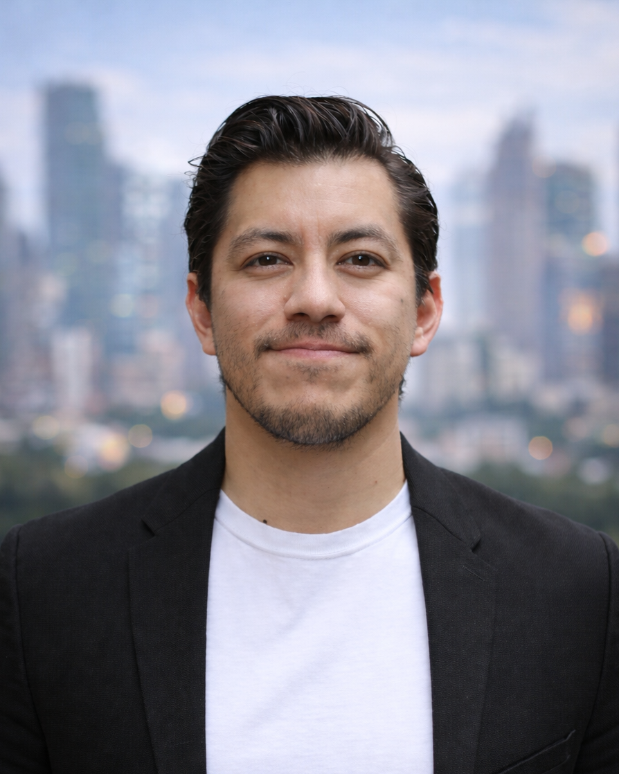 Joven hombre con cabello oscuro y barba, vistiendo chaqueta negra y camiseta blanca, frente a un fondo urbano con edificios y cielo