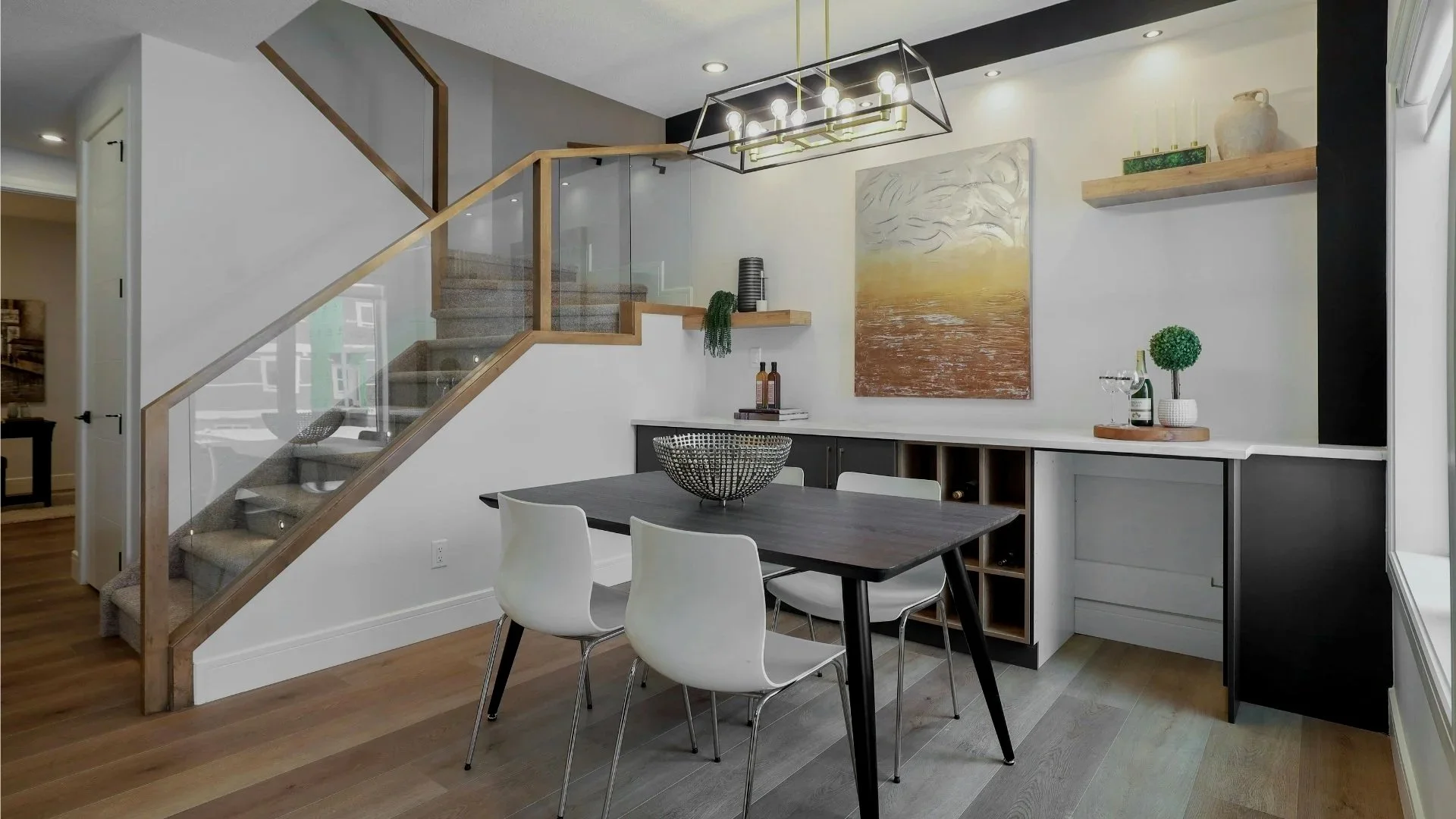 Interior de comedor con mesa, sillas blancas, escalera de madera con barandilla de vidrio, decoración moderna, cuadro en la pared y estantería con objetos decorativos.