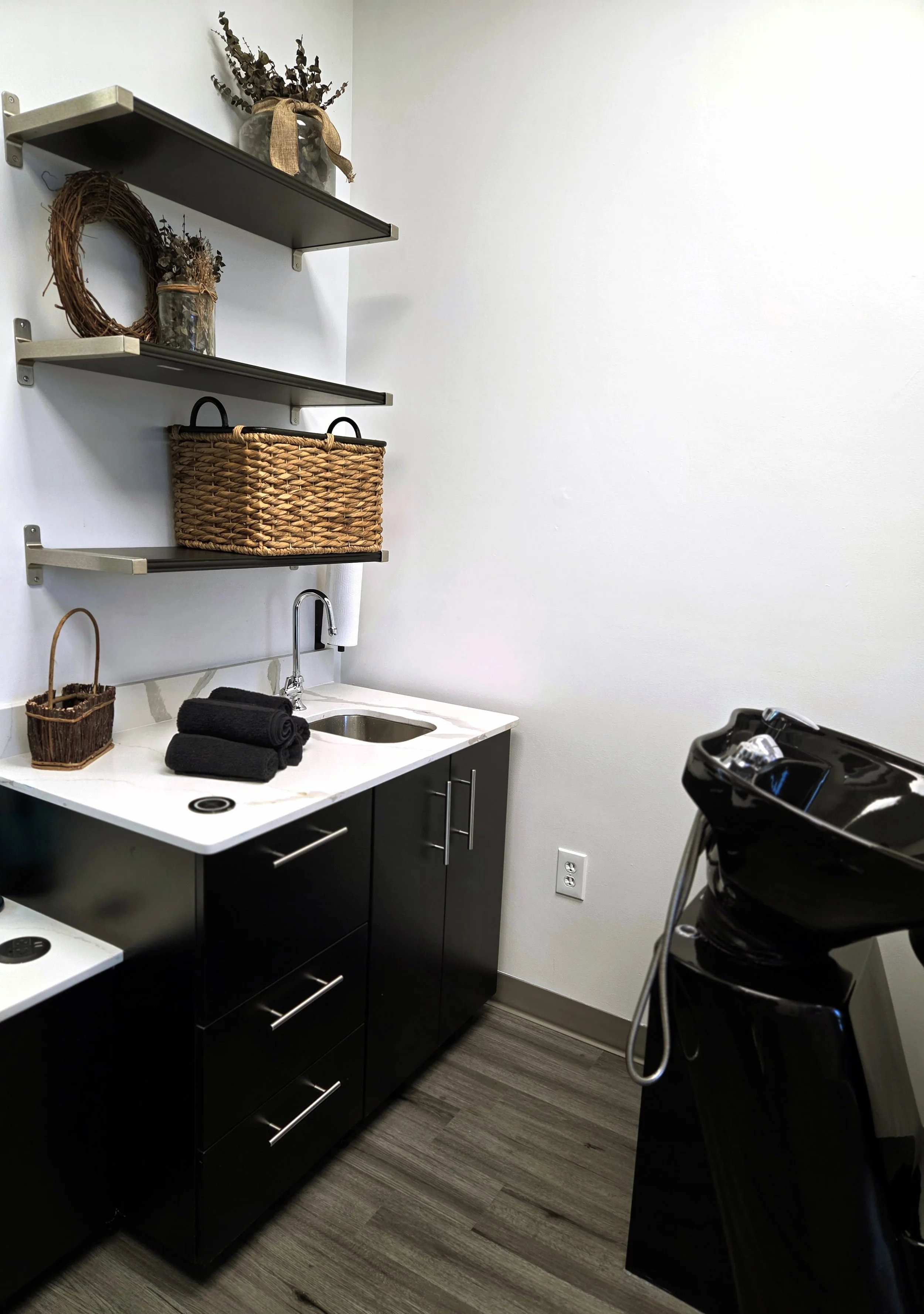 A salon or grooming station with a black sink and countertop, black cabinets, shelves with decorations including a wreath and jars with dried flowers, black towels, and a black hair washing station in the foreground.
