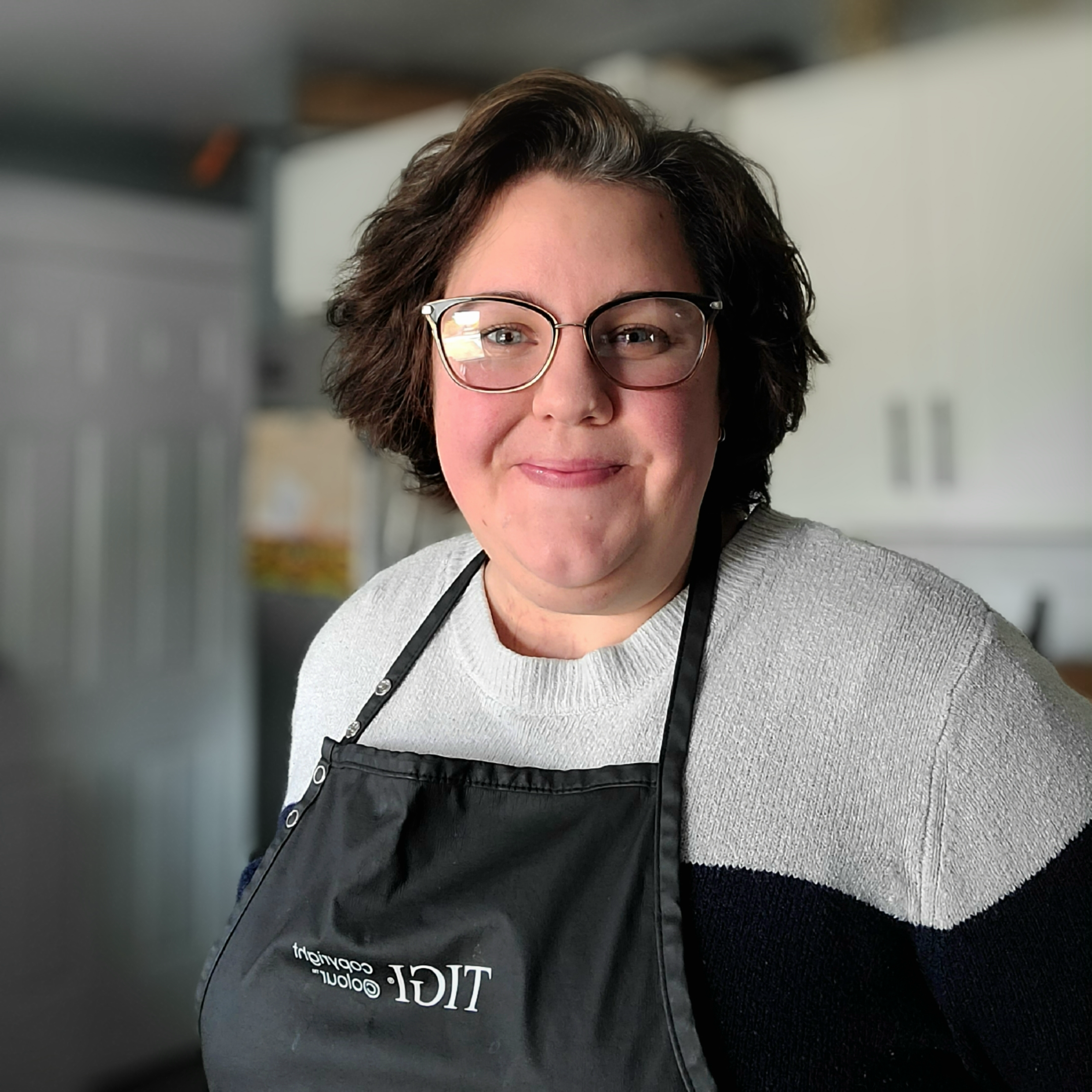 Close-up of a woman with short brown hair and glasses wearing a gray sweater and a black apron, smiling at the camera, in a kitchen setting.