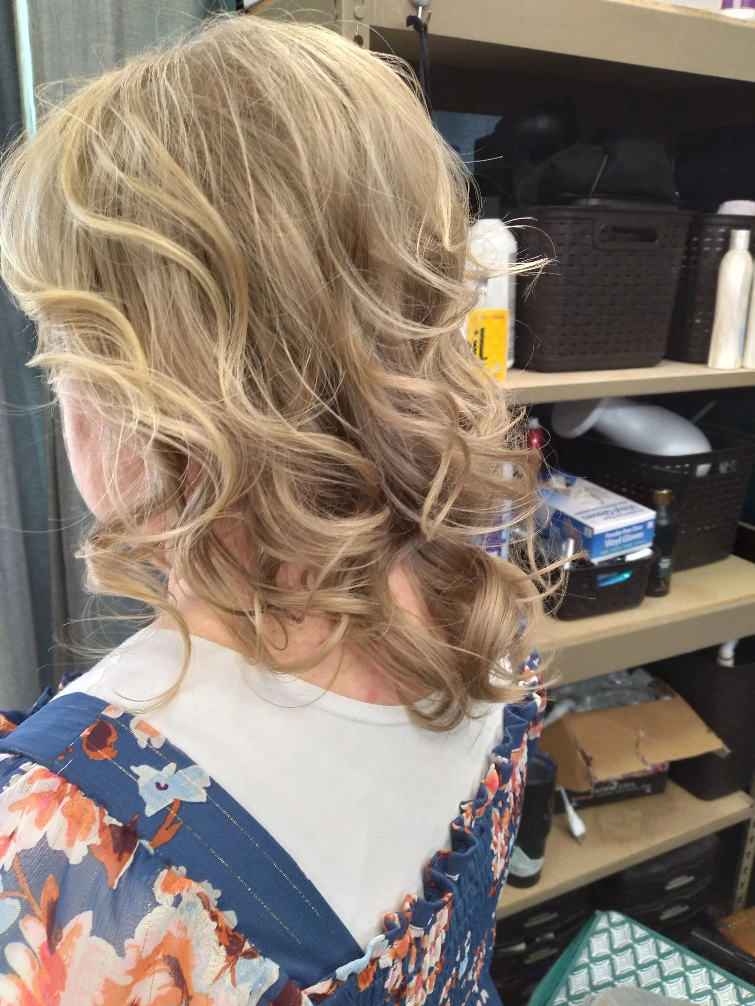 A woman with blonde, curly hair wearing a floral-patterned top and a white shirt underneath, standing in front of shelves with various objects, including a black basket, bottles, and boxes.