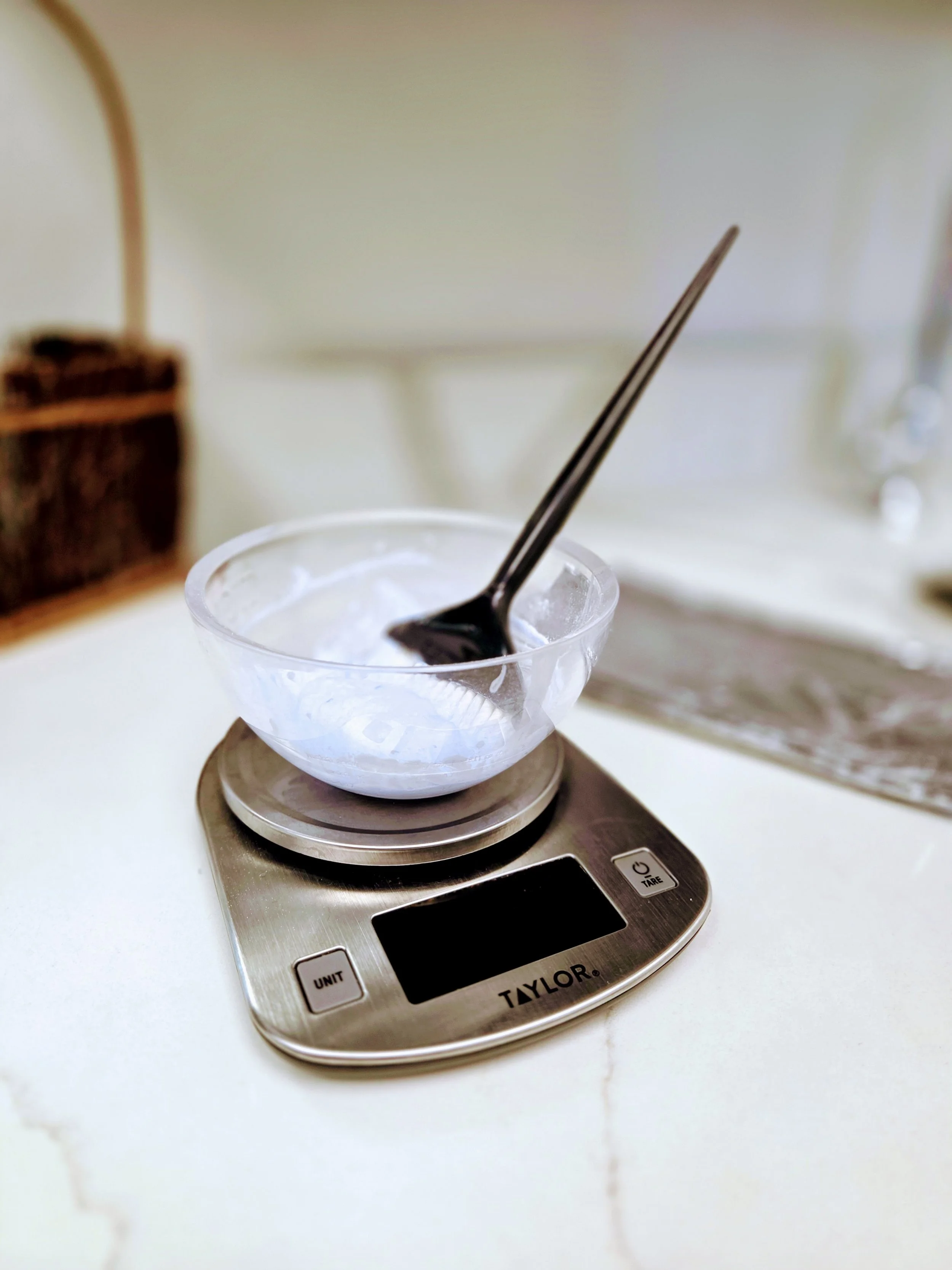 A digital kitchen scale with a small glass bowl on top, containing a dark substance and a pair of metal tongs. The scale displays the brand 'TAYLOR' and has buttons labeled 'UNIT' and 'TARE'.