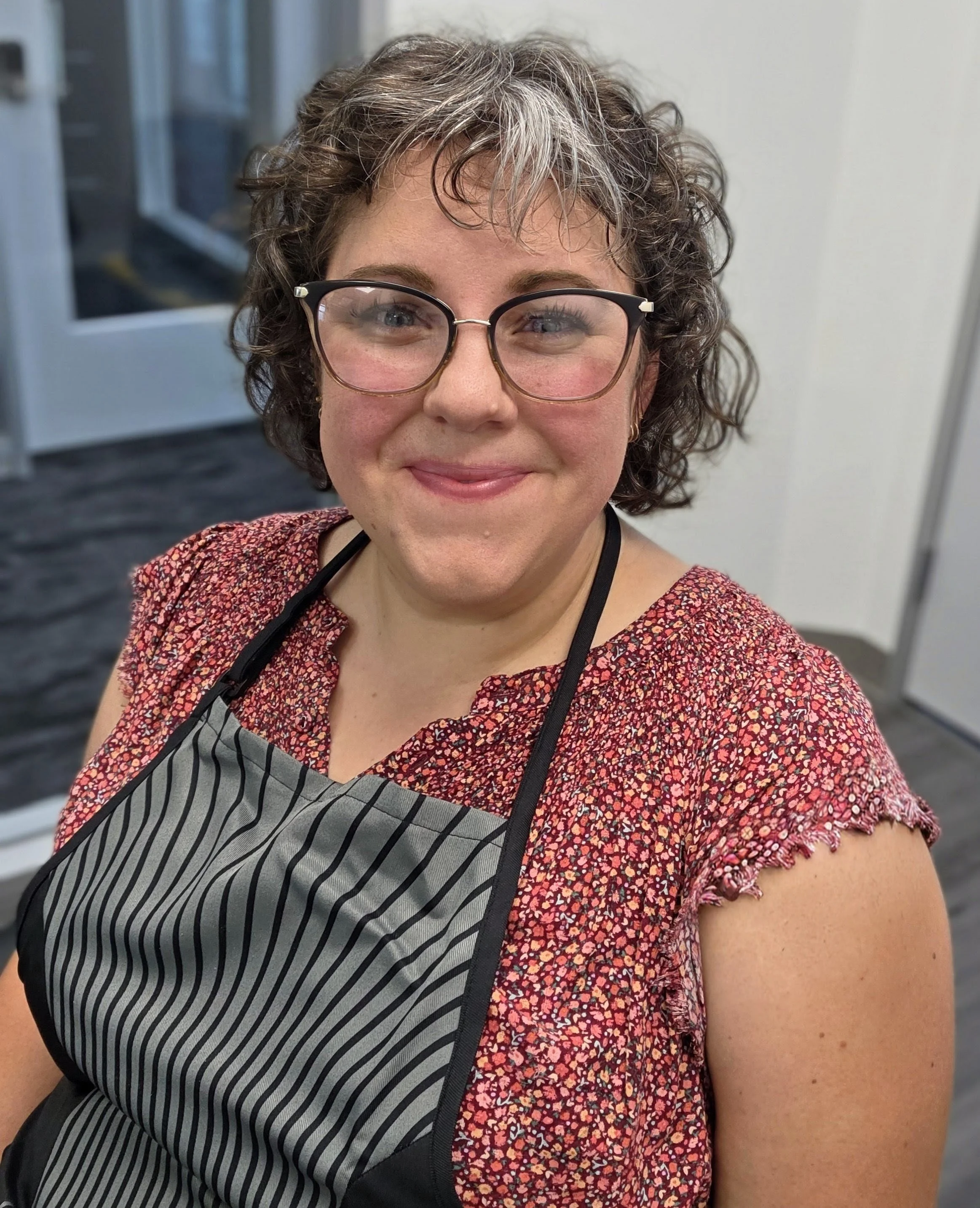 A woman with curly hair and glasses smiling for a selfie in an indoor setting, wearing a floral top and a black and white striped apron.