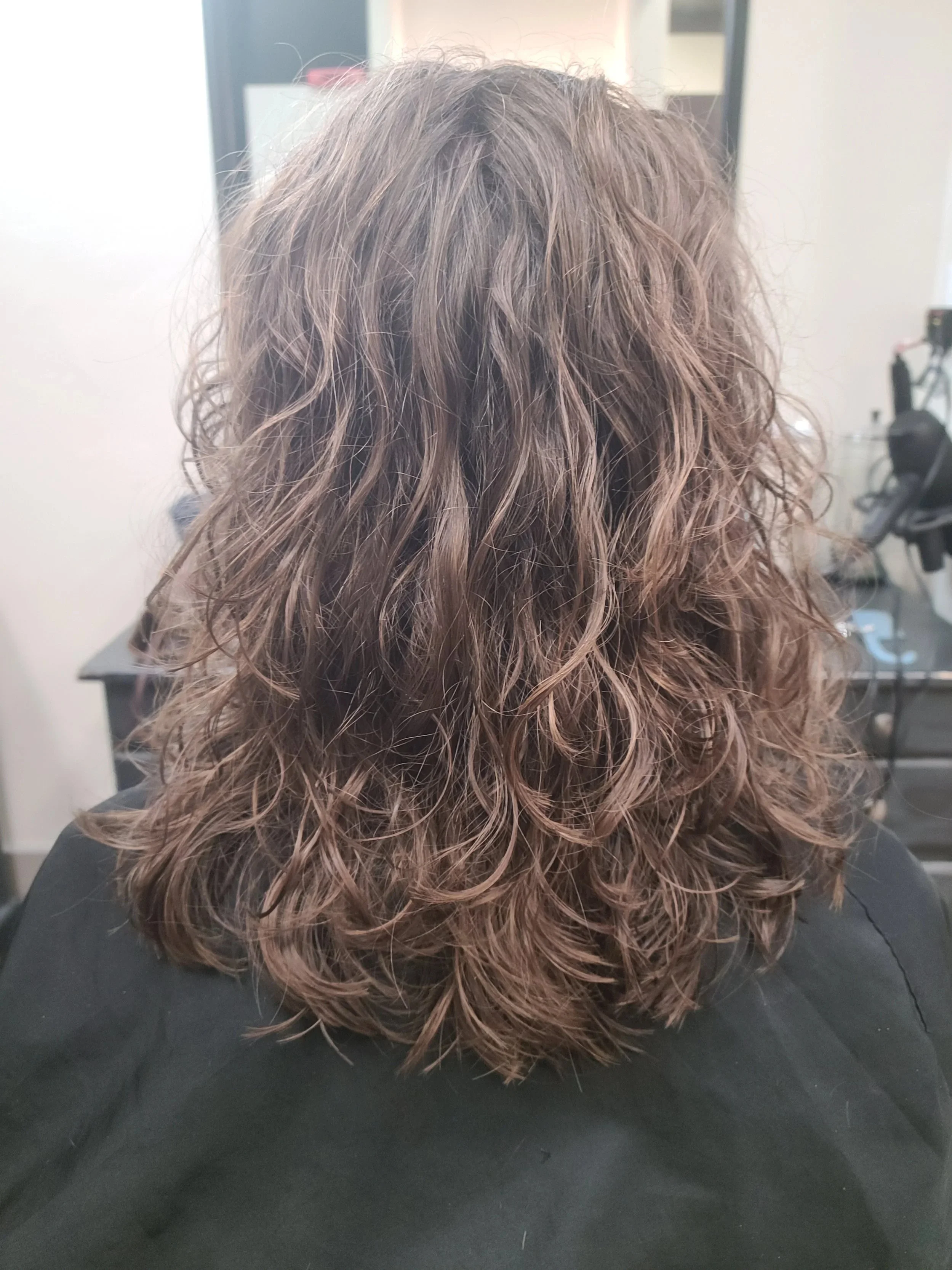 Back view of shoulder-length, curly hair with blonde and light brown highlights, resting on a black surface in a salon setting.
