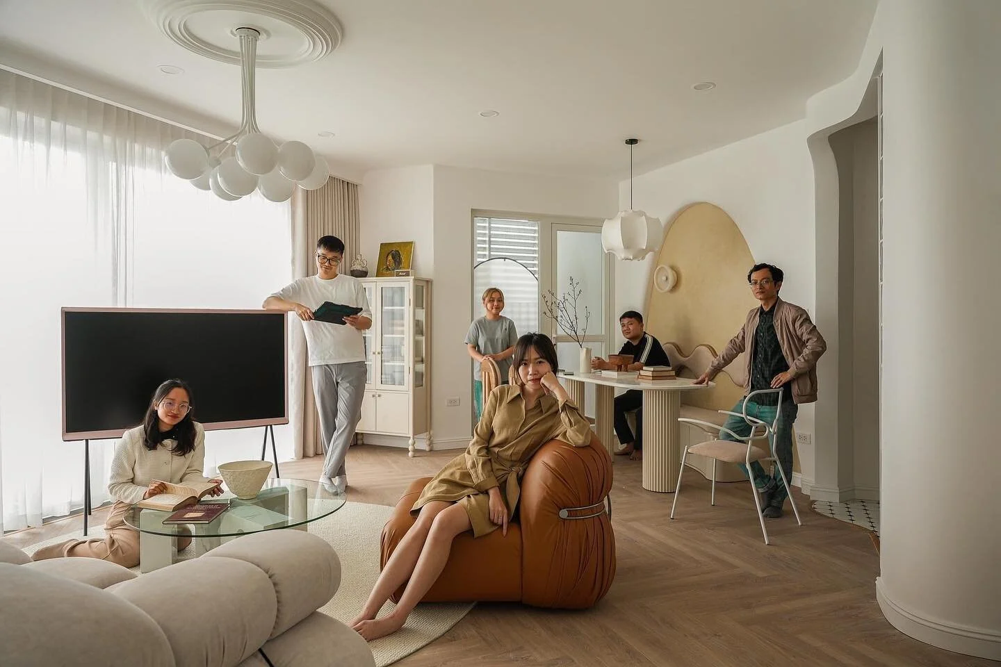 A group of six people in a bright, modern living room with white walls and wood flooring. Some are sitting on a light-colored sofa and a leather bean bag, while others stand near a white table and a TV. The room has large windows with sheer curtains, and contemporary light fixtures hang from the ceiling.