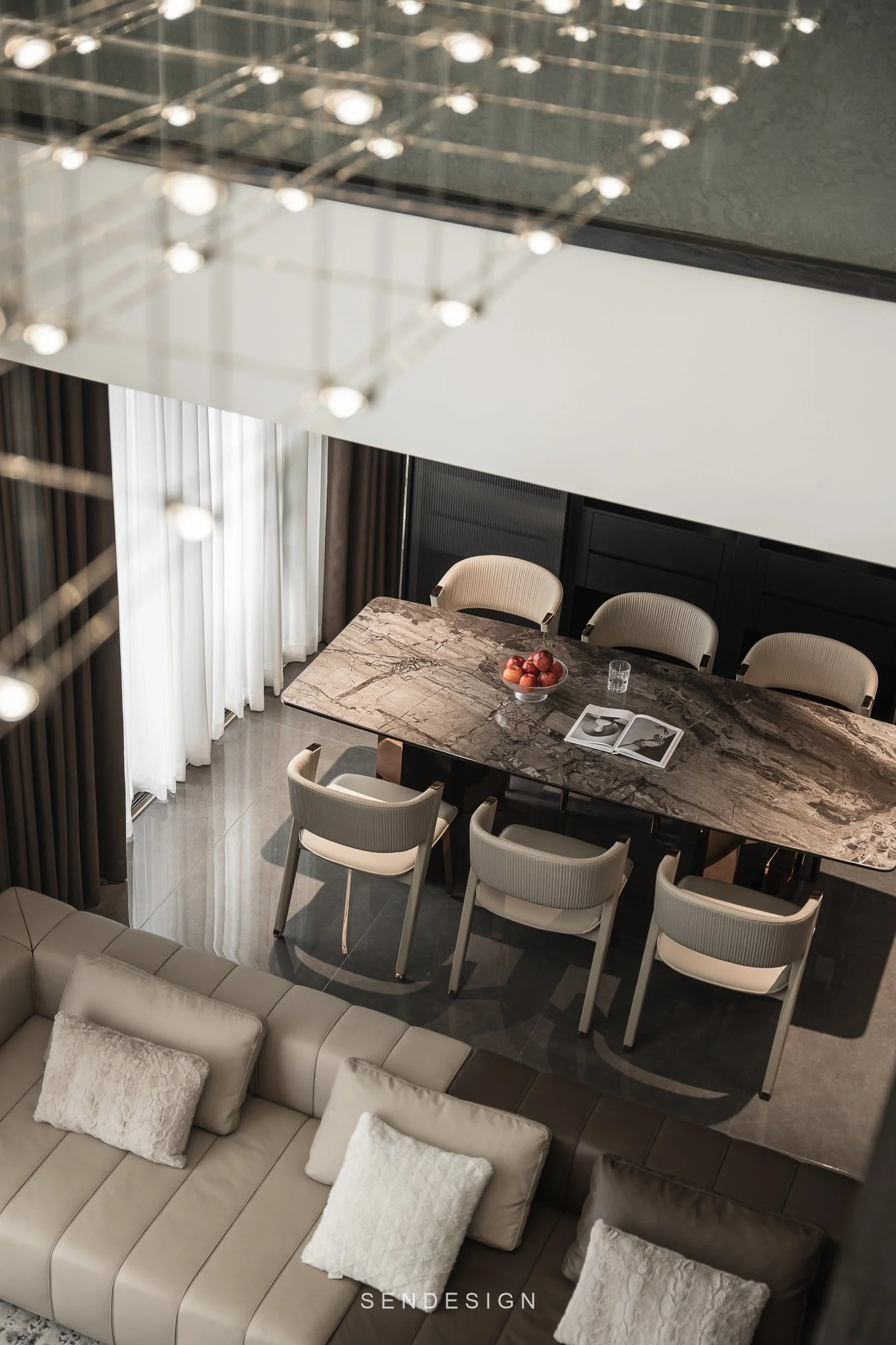 A view of a modern dining room with a large marble table surrounded by seven beige chairs, a bowl of apples on the table, and a magazine. Light curtains hang over the windows in the background, and a beige sectional sofa with pillows is in the foregr
